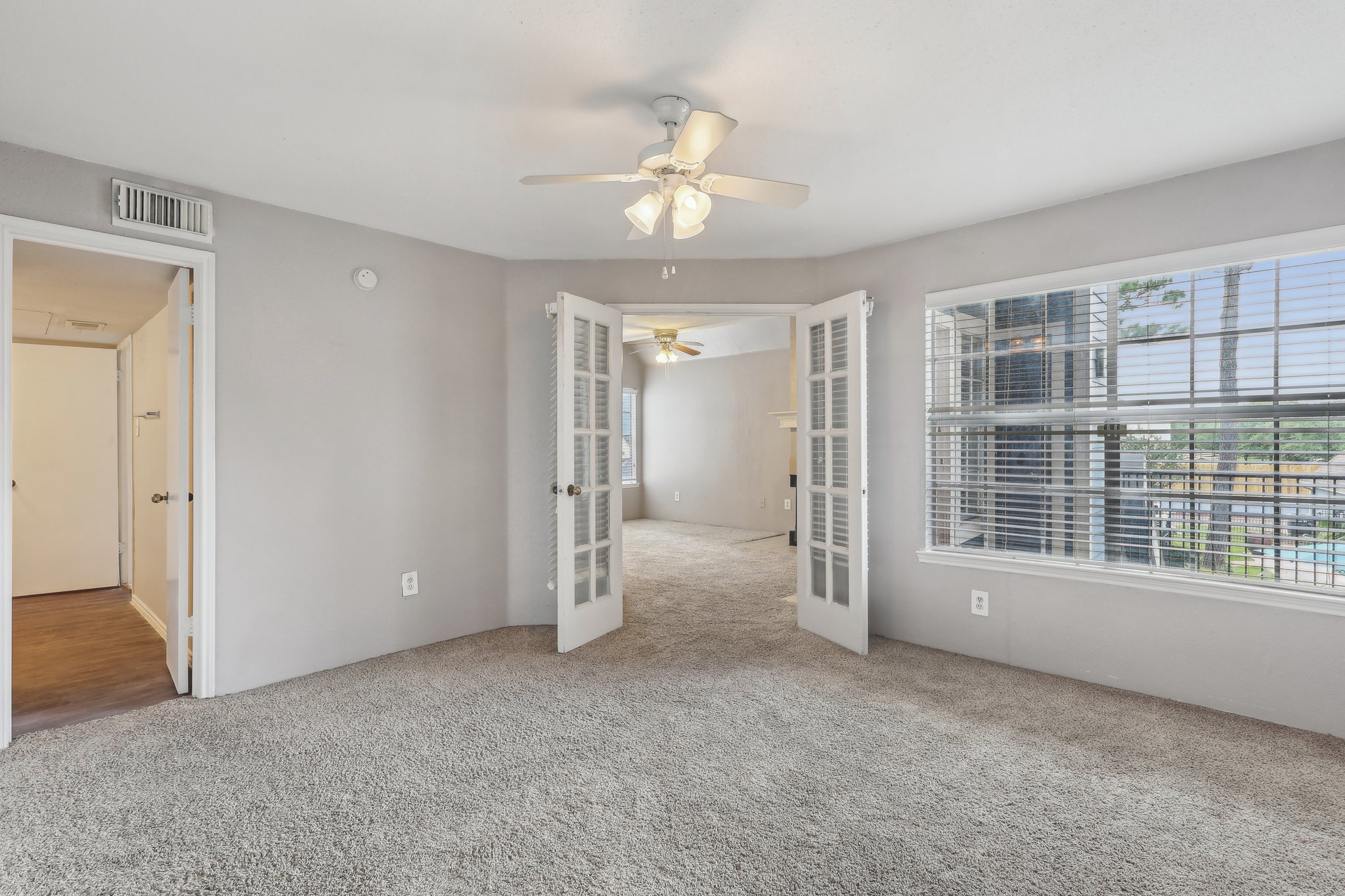 Spacious living area with beige carpet, featuring two ceiling fans and large windows providing natural light. The room includes French doors leading to another space, creating an open and airy atmosphere. Light gray walls complement the neutral decor.