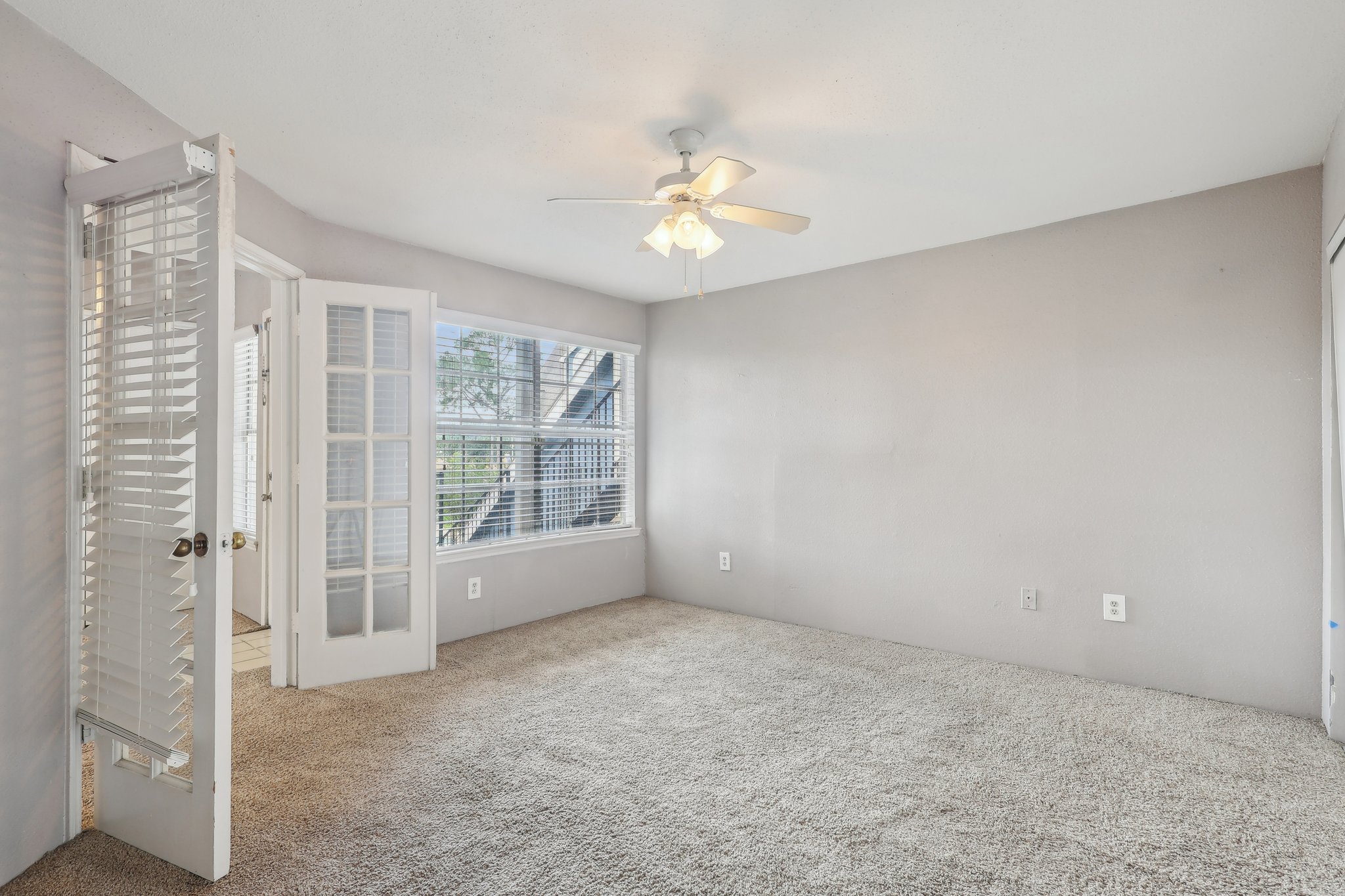 A vacant room with light gray walls and carpet. It features a ceiling fan, a large window with blinds allowing natural light, and an adjoining door leading to another space. The room appears spacious and tidy.