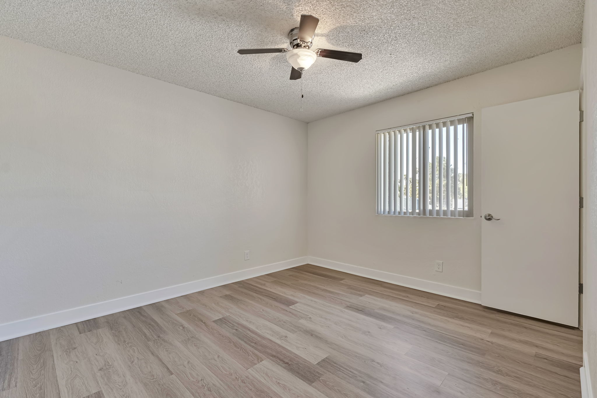 A well-lit empty room with light-colored walls and a ceiling fan. It features wooden laminate flooring, a vertically-slatted window with natural light coming through, and a closed white door on one side. The space is clean and ready for furnishing, giving a fresh and airy feel.