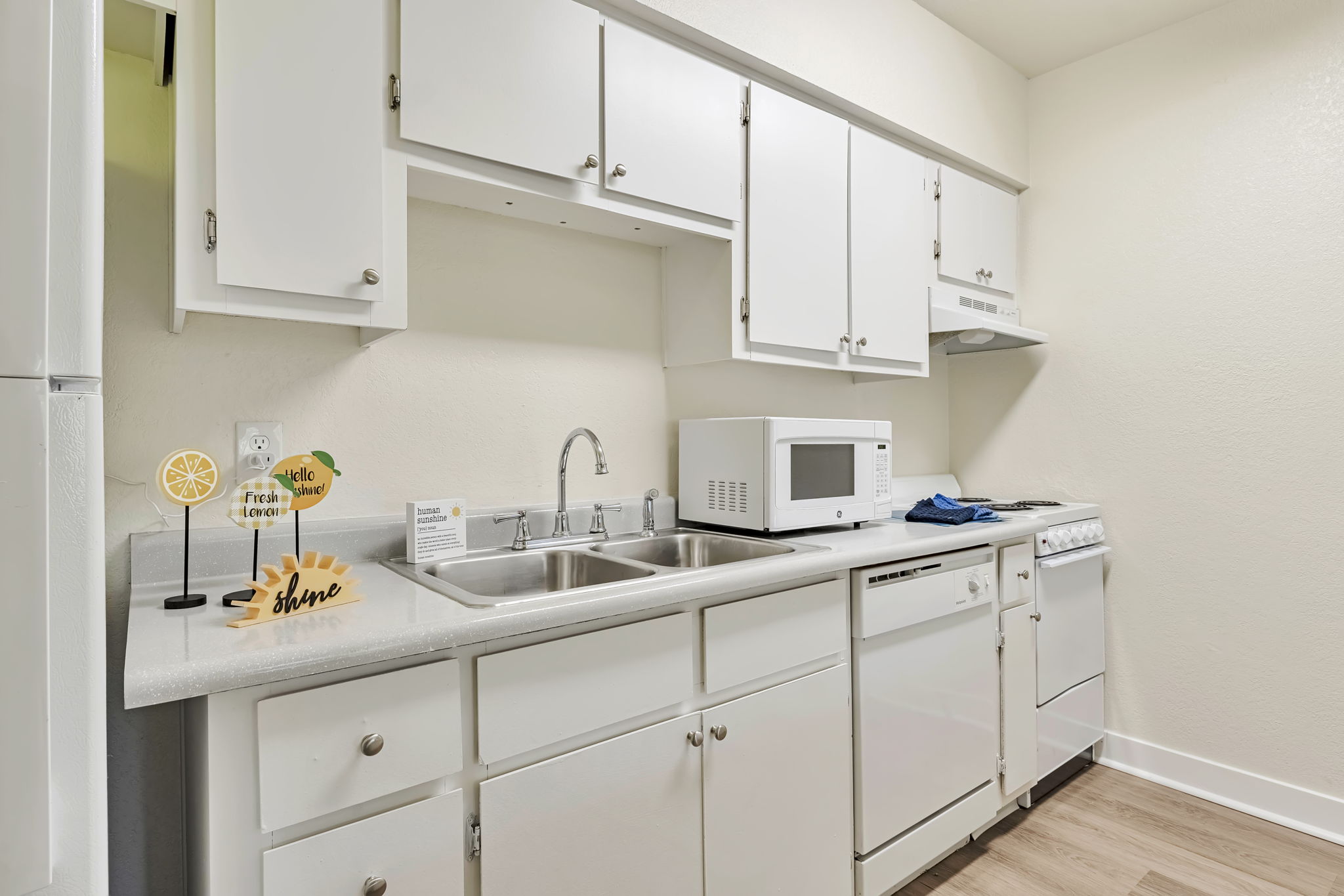 A clean, modern kitchen featuring white cabinets, a stainless steel sink, a microwave, and a stove. There's a small decorative display with a lemon slice and the word "hello," adding a cheerful touch to the space. The countertops are light-colored, and the flooring is laminate.