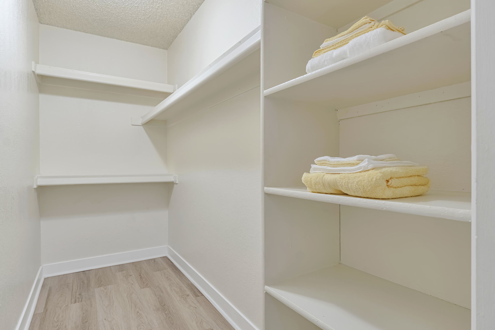 A clean and organized closet with two shelves on either side. The left side has empty shelves, while the right side features neatly folded yellow and white towels stacked on the lower shelf. The walls are painted white, and the floor has a light wooden finish.