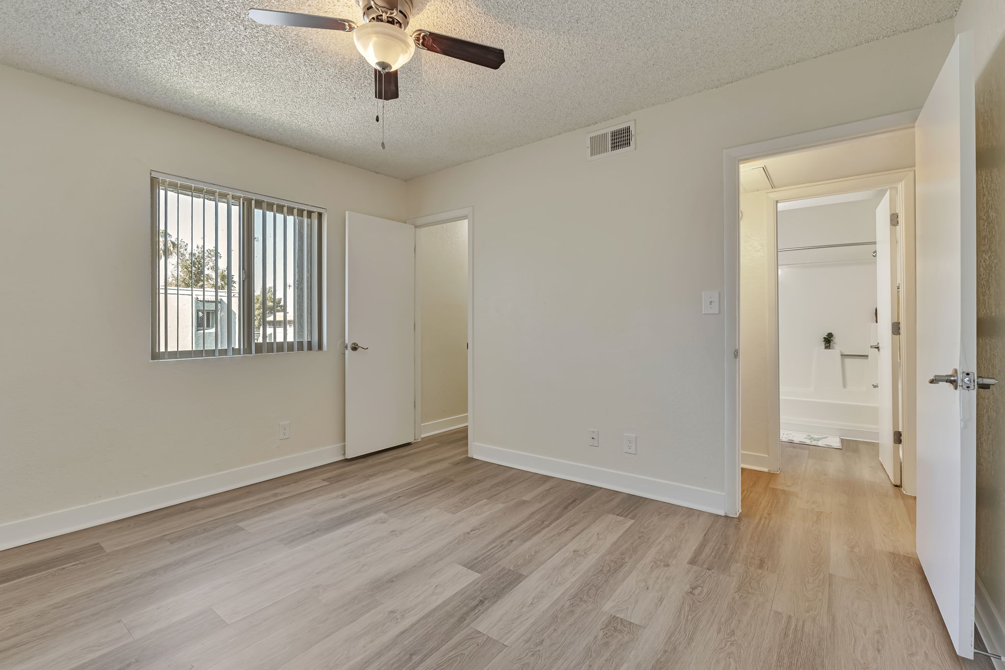 A bright, empty room featuring light-colored walls, a ceiling fan, and laminate flooring. Natural light comes through a window with vertical blinds. A doorway leads to a bathroom, visible in the background, while another door is slightly ajar, suggesting access to another room or space.