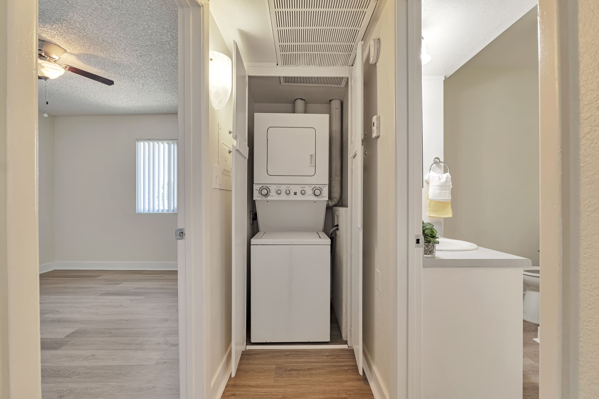 A view of a small laundry area featuring a stacked washer and dryer, situated between two doorways. One doorway leads to a bathroom with a towel hanging, and the other leads to a living space with a ceiling fan and vertical blinds. The flooring is a light wood color.