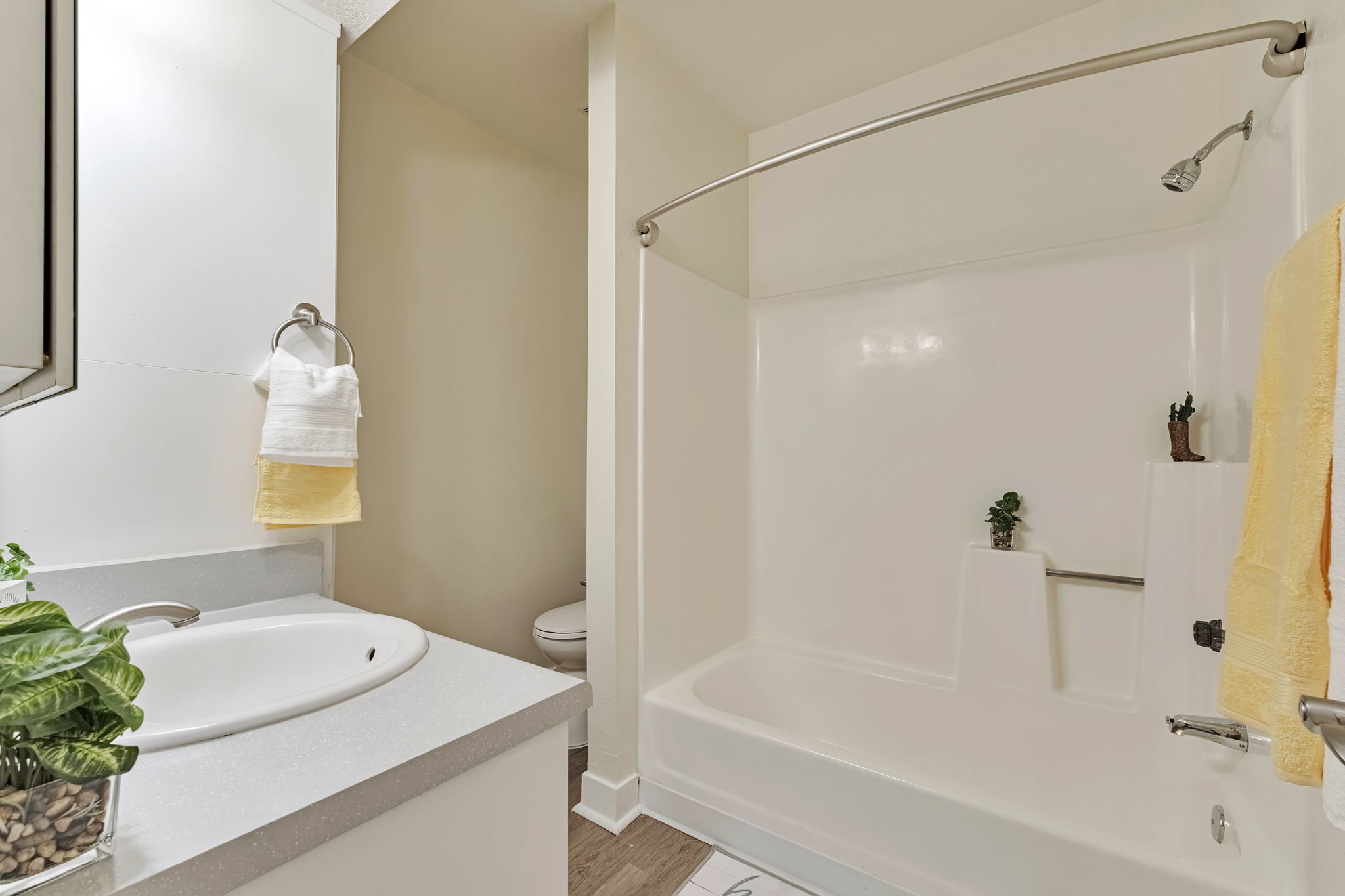 A clean and modern bathroom featuring a bathtub with a shower rod, a white sink with a marble countertop, and a small potted plant. Soft yellow towels hang from a rack, and a toilet is visible in the background. The walls are painted a light color, creating a bright and airy atmosphere.