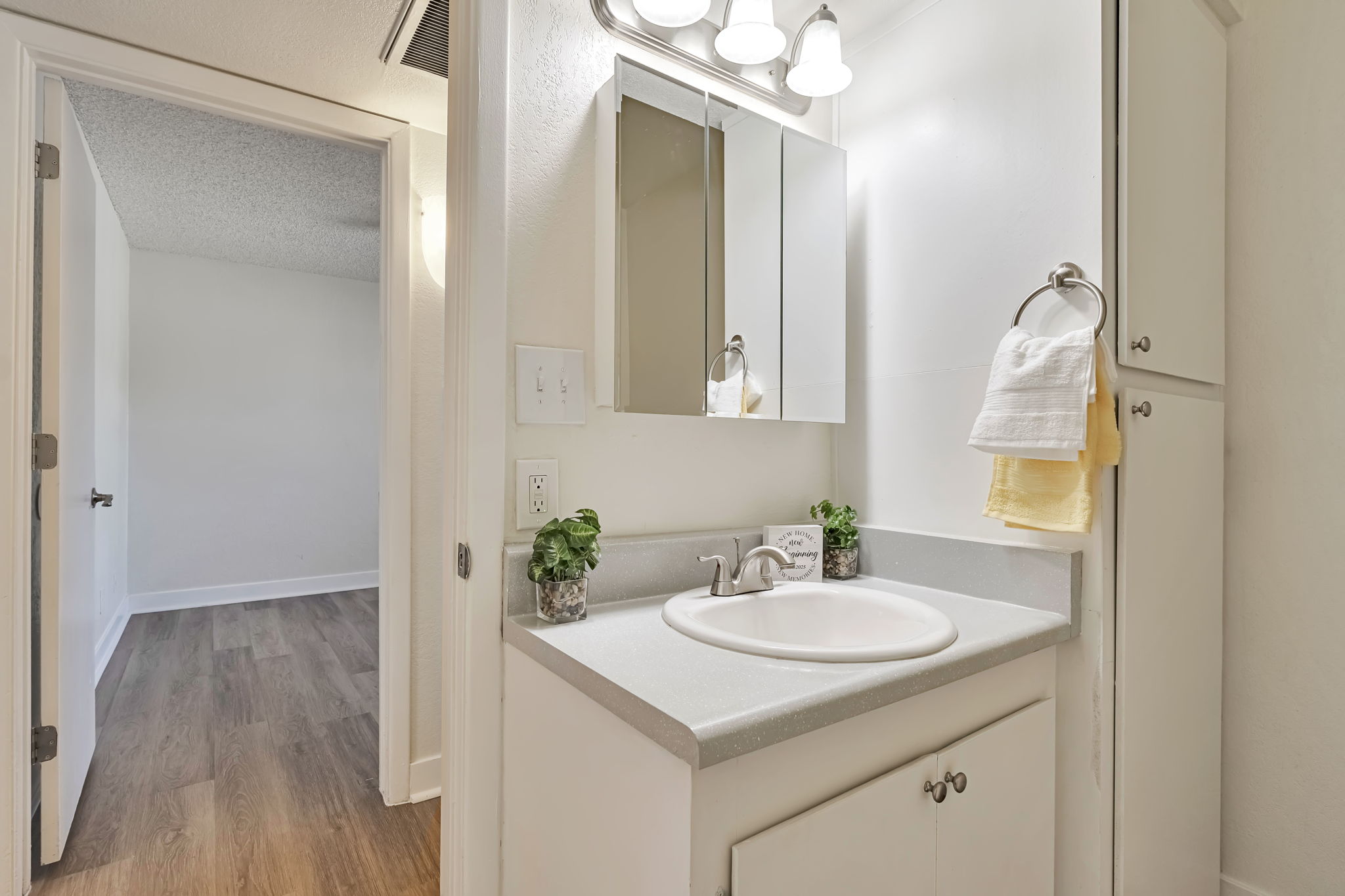 A clean and modern bathroom featuring a white sink with a light gray countertop, wall-mounted mirrors, and neatly arranged towels. Small potted plants add a touch of greenery. The doorway in the background leads to a light-colored room with wooden flooring.
