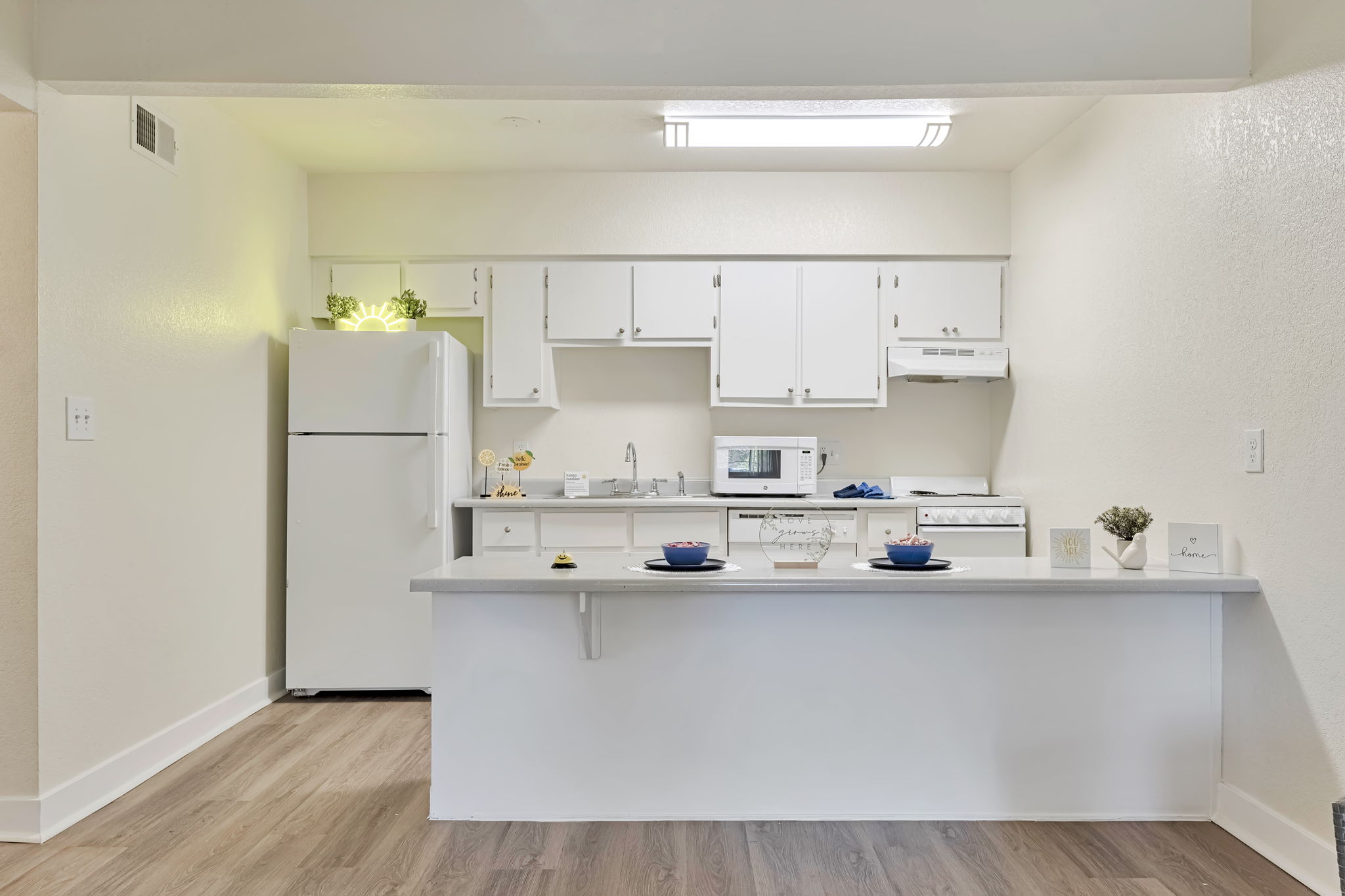 A bright, modern kitchen featuring white cabinetry, a refrigerator, a microwave, and a stove. A countertop with two blue plates and small decor adds a cozy touch. Natural light enhances the space, creating a welcoming atmosphere with wooden flooring and minimalist design elements.