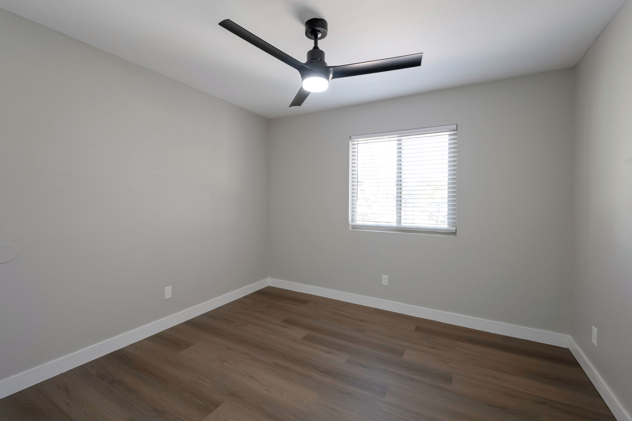 A minimalist bedroom with light gray walls, a ceiling fan with a light fixture, and a window covered with white blinds. The floor is made of light brown wood, and the room is empty, featuring clean lines and a bright, airy feel.