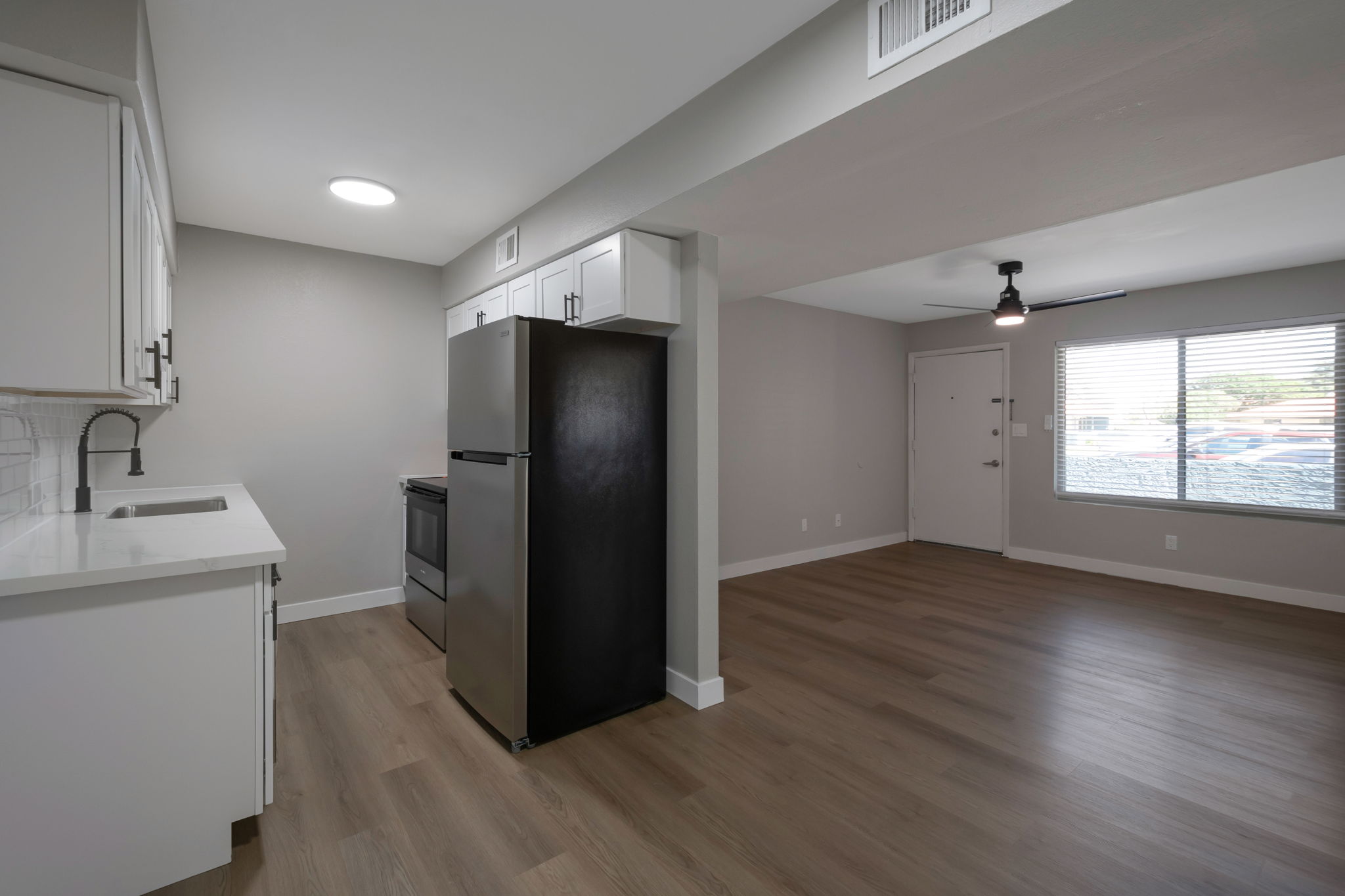 Interior view of a modern kitchen and living area. The kitchen features white cabinetry, a stainless steel sink, and black appliances. The living area has a large window with blinds, offering natural light, and includes light-colored flooring. The walls are painted in a neutral tone, creating a spacious and inviting atmosphere.