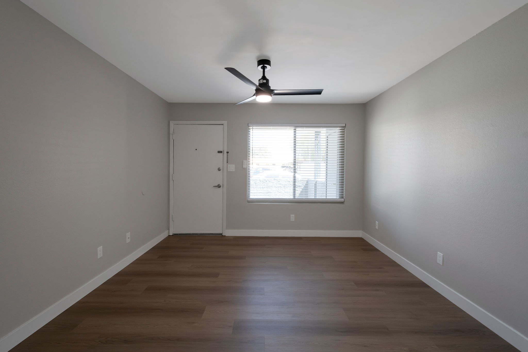 A well-lit empty room featuring light gray walls and a wooden floor. There's a ceiling fan with a light fixture, a closed door on the left, and a window with blinds on the right allowing natural light to enter. The overall ambiance is simple and modern, suitable for various purposes.
