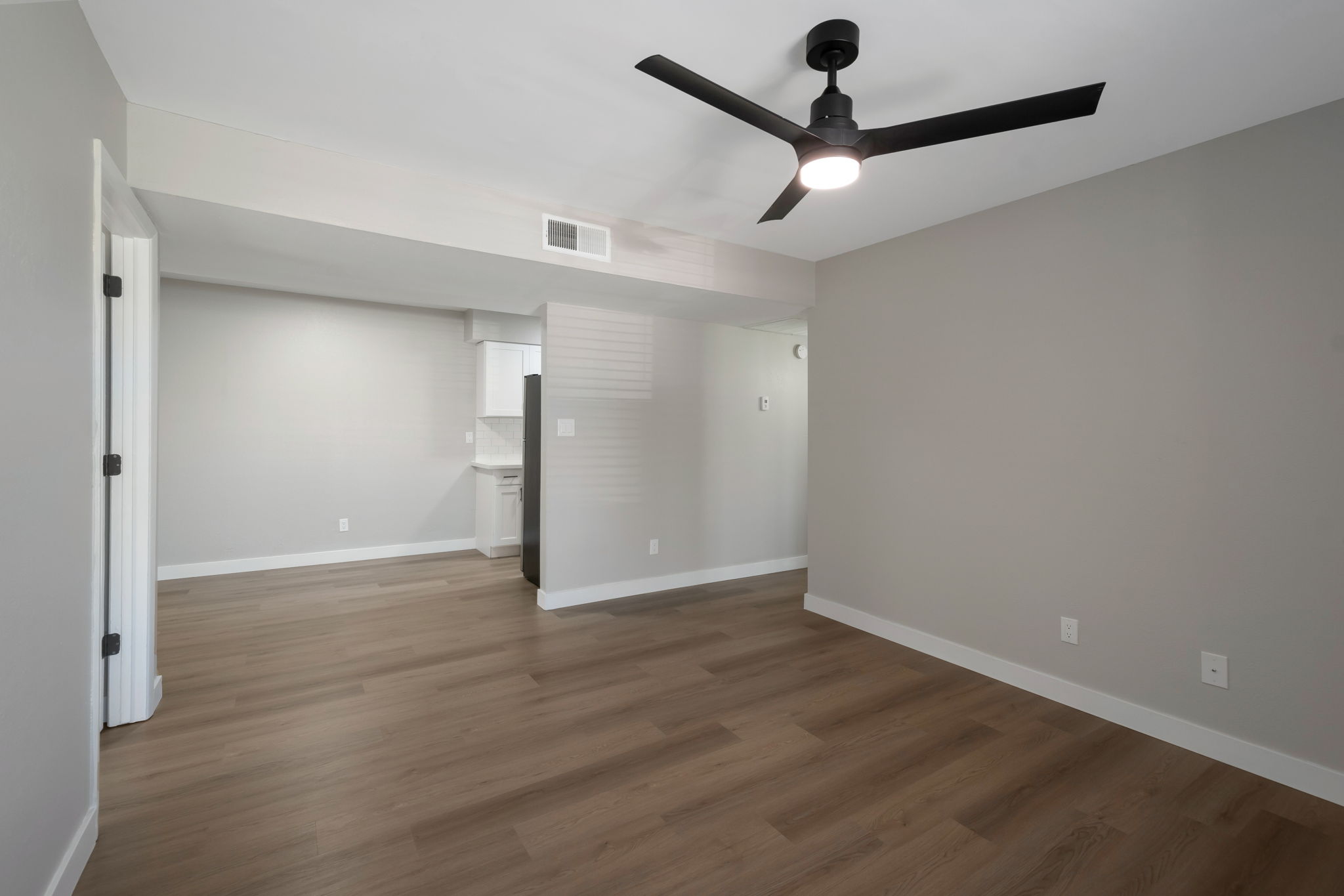 A spacious, empty living area featuring light gray walls and a modern ceiling fan. The floor is covered with light wood laminate, and there's an open doorway leading to a kitchenette in the background. Natural light floods the room, creating a bright and airy atmosphere.