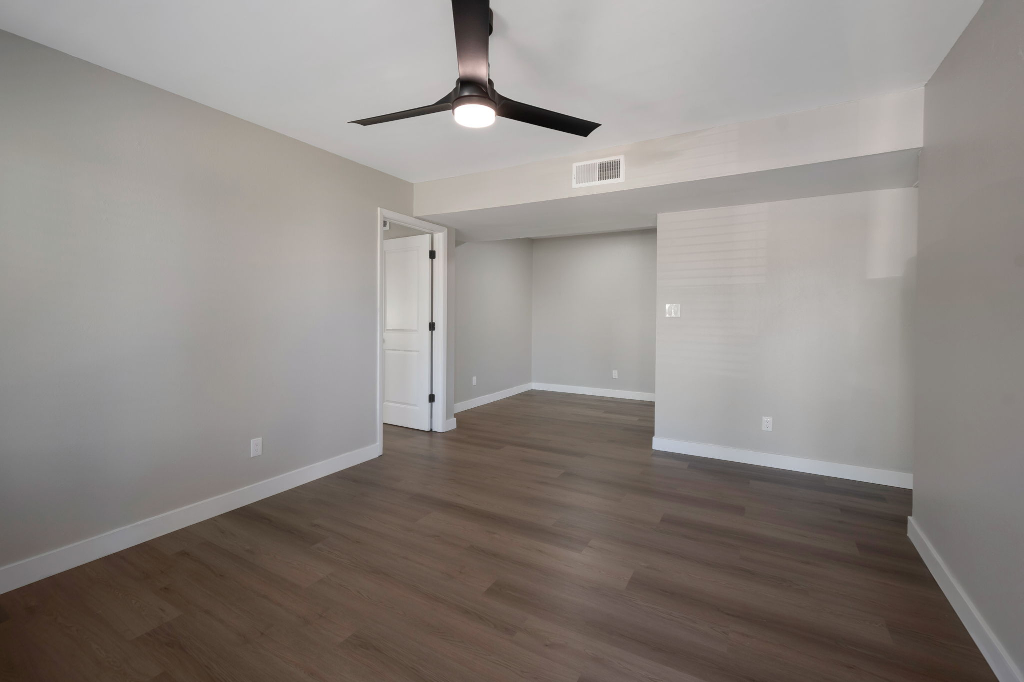 Empty room with light gray walls and wooden flooring. A ceiling fan is mounted on the ceiling. There is a closed white door on the left side and a small wall-mounted fixture visible in the back corner. The room is well-lit with natural light coming through the windows.