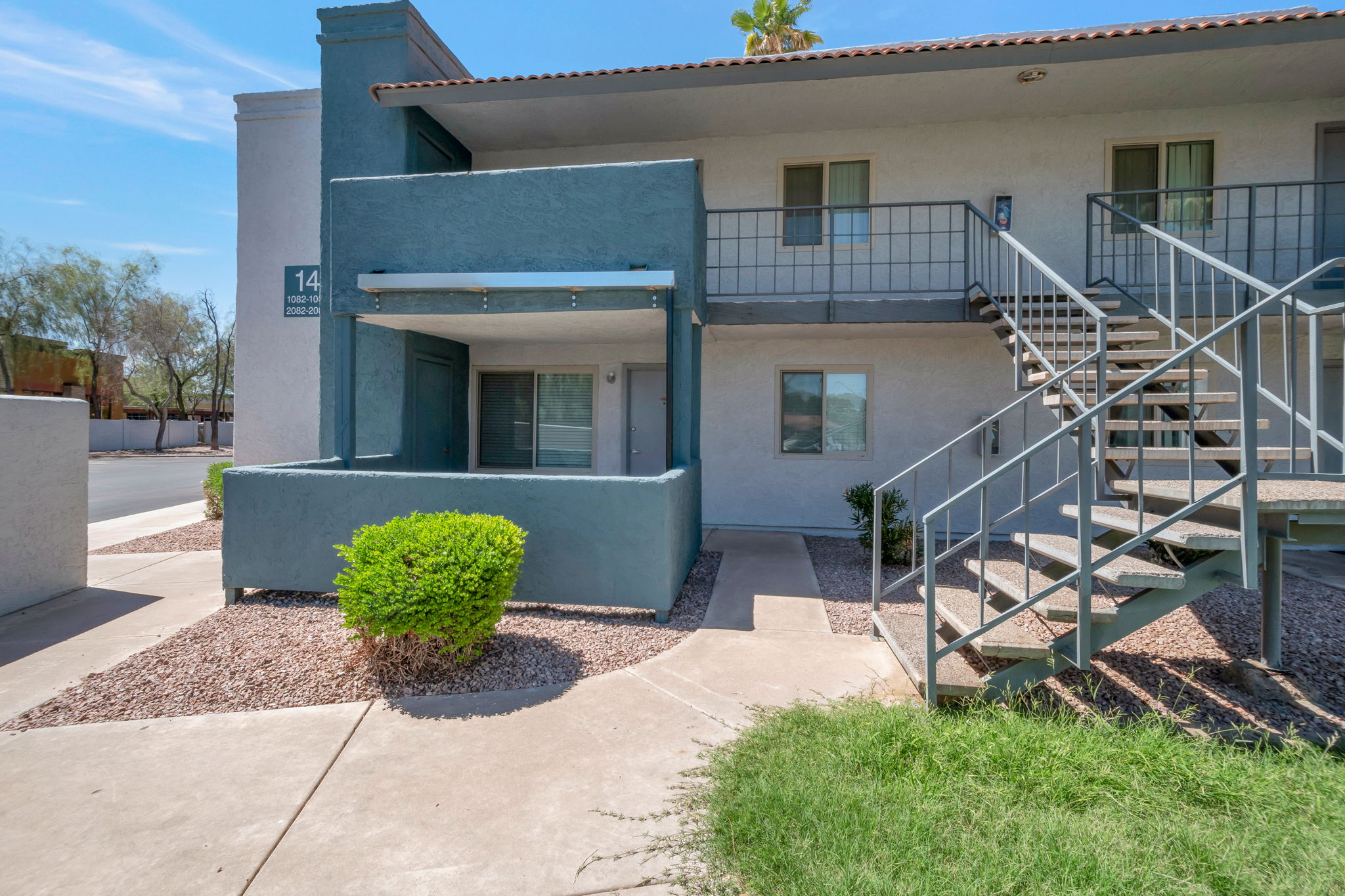 A blue-gray apartment building with a staircase leading to the second floor. The ground level features a small patio area with a railing. Landscaping includes a bush and gravel surrounding the walkway. The scene has clear blue skies and is set in a residential neighborhood.
