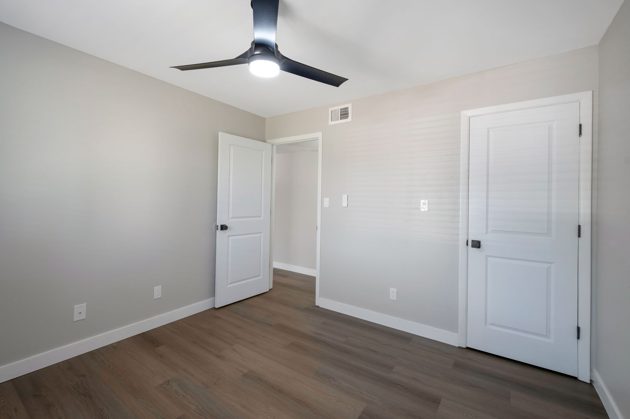 Empty room with light gray walls and a wooden floor. A ceiling fan is mounted in the center, providing overhead lighting. There are two white doors, one leading to a closet or another room, and the other is a standard door. The space is bright and minimalistic, ideal for personal customization.