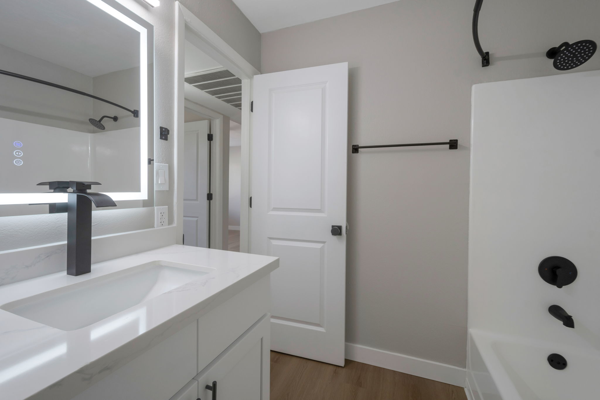 Modern bathroom featuring a clean, minimalist design. It includes a white vanity with a rectangular sink and a sleek, black faucet. A backlit mirror is mounted above the sink. The walls are painted in a soft gray, and there is a shower area with a black showerhead. A door leads to another room, and a towel rack is visible.