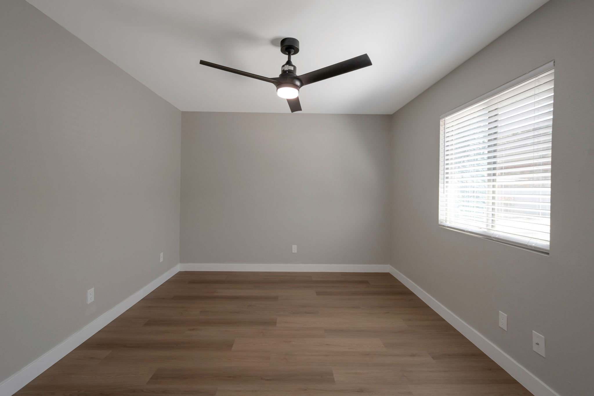 A spacious, empty room featuring light gray walls and light wood flooring. A modern ceiling fan with a light is mounted in the center. A window with white blinds allows natural light to enter, illuminating the room's minimalistic design.