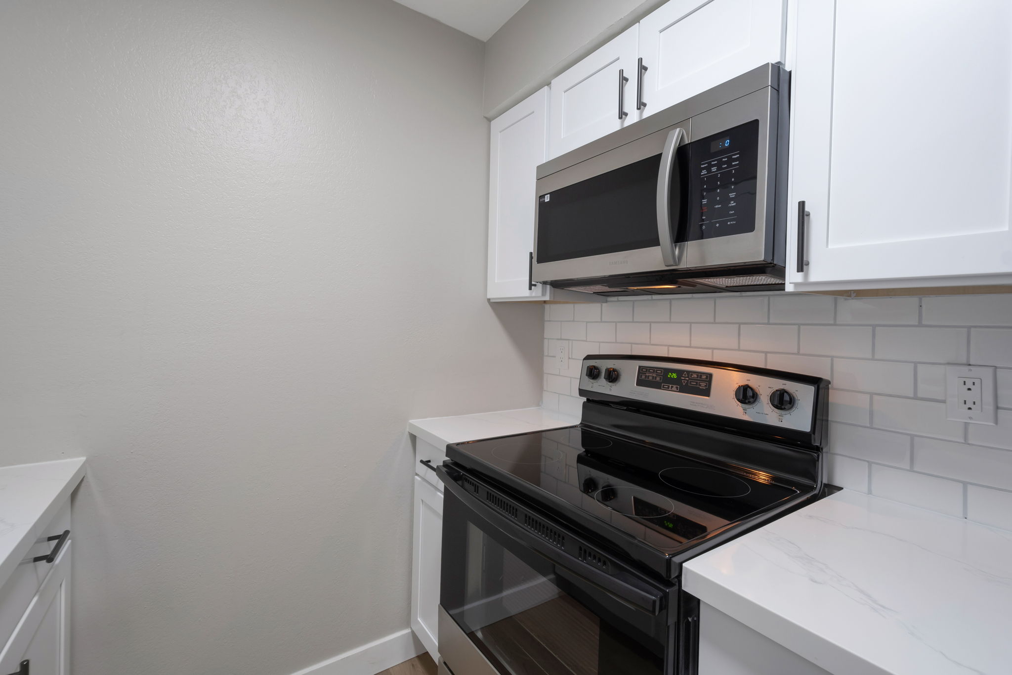 Modern kitchen featuring white cabinetry, a stainless steel microwave mounted above an electric stove, and a black countertop. The backsplash consists of white subway tiles, and the overall color scheme is light and contemporary. The space appears clean and well-organized.