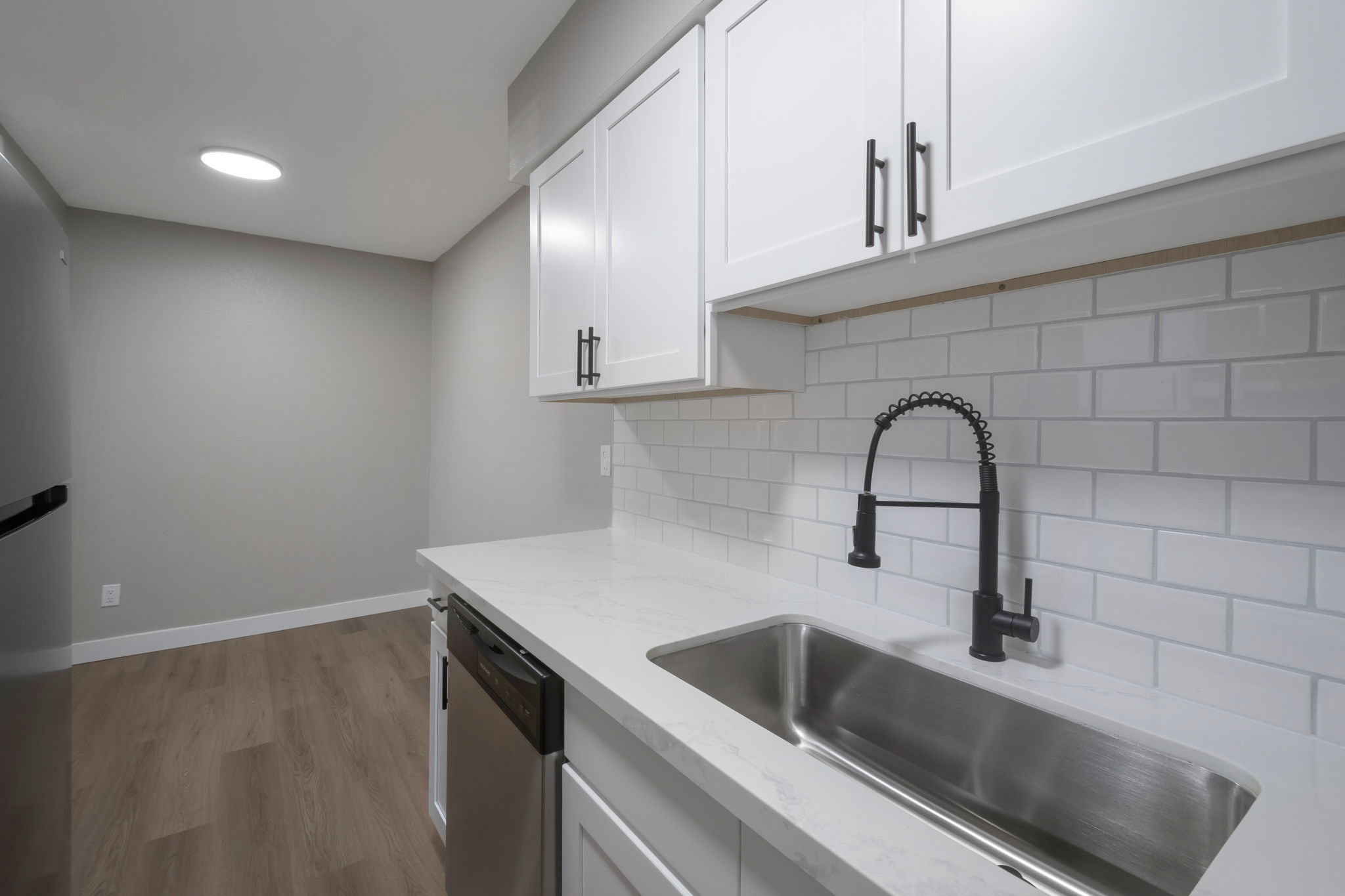Modern kitchen with white cabinets, a stainless steel sink with a black pull-down faucet, and a dishwasher. Features a light-colored countertop and white subway tile backsplash. The space has neutral walls and a wooden floor, with a gray refrigerator visible in the corner. Bright, well-lit environment.
