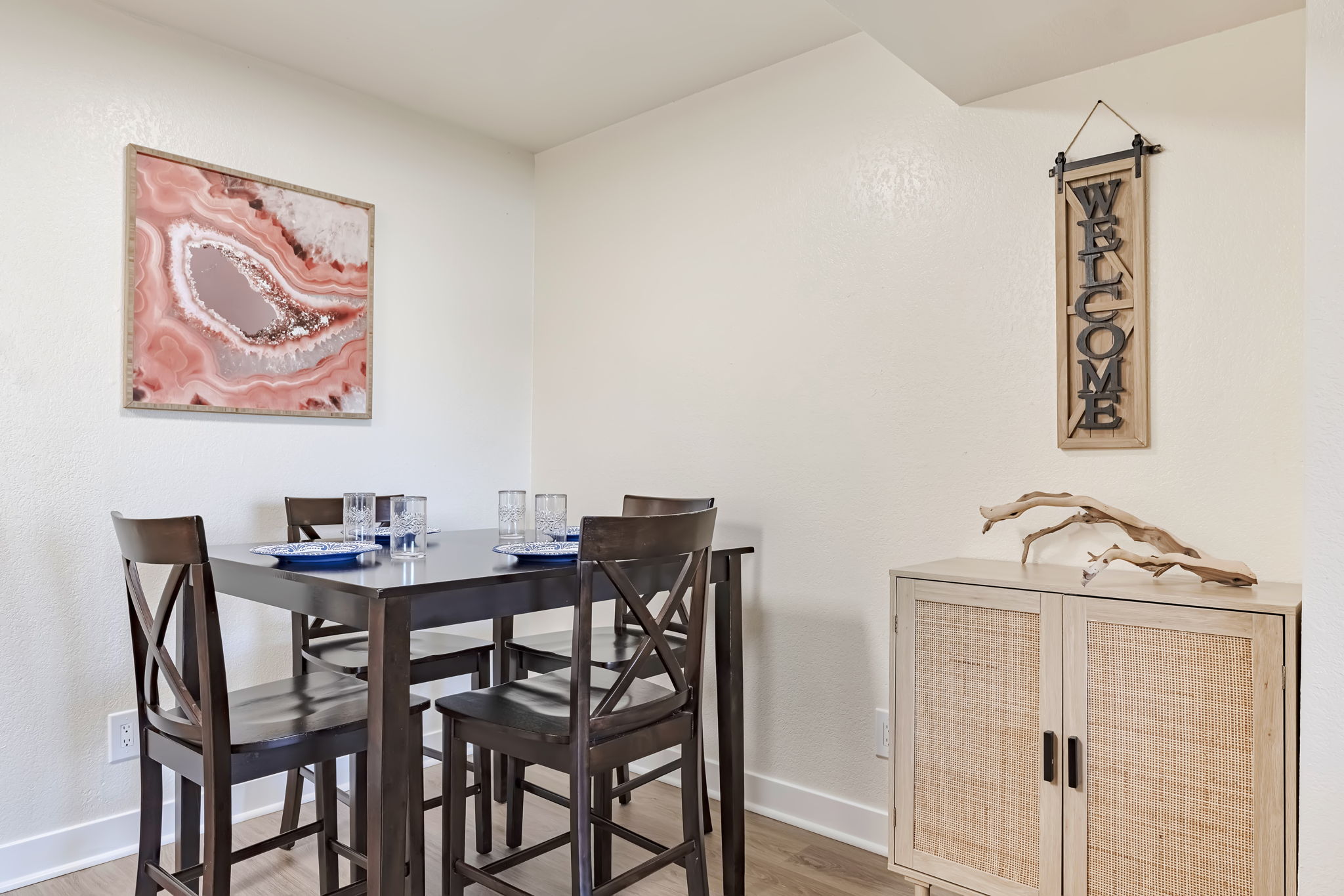 A dining area featuring a dark wood table with matching chairs, set for four with glasses and blue plates. A pink and white abstract painting hangs on the wall, and a wooden "WELCOME" sign is displayed above a light-colored cabinet with woven accents. The space is bright and modern.