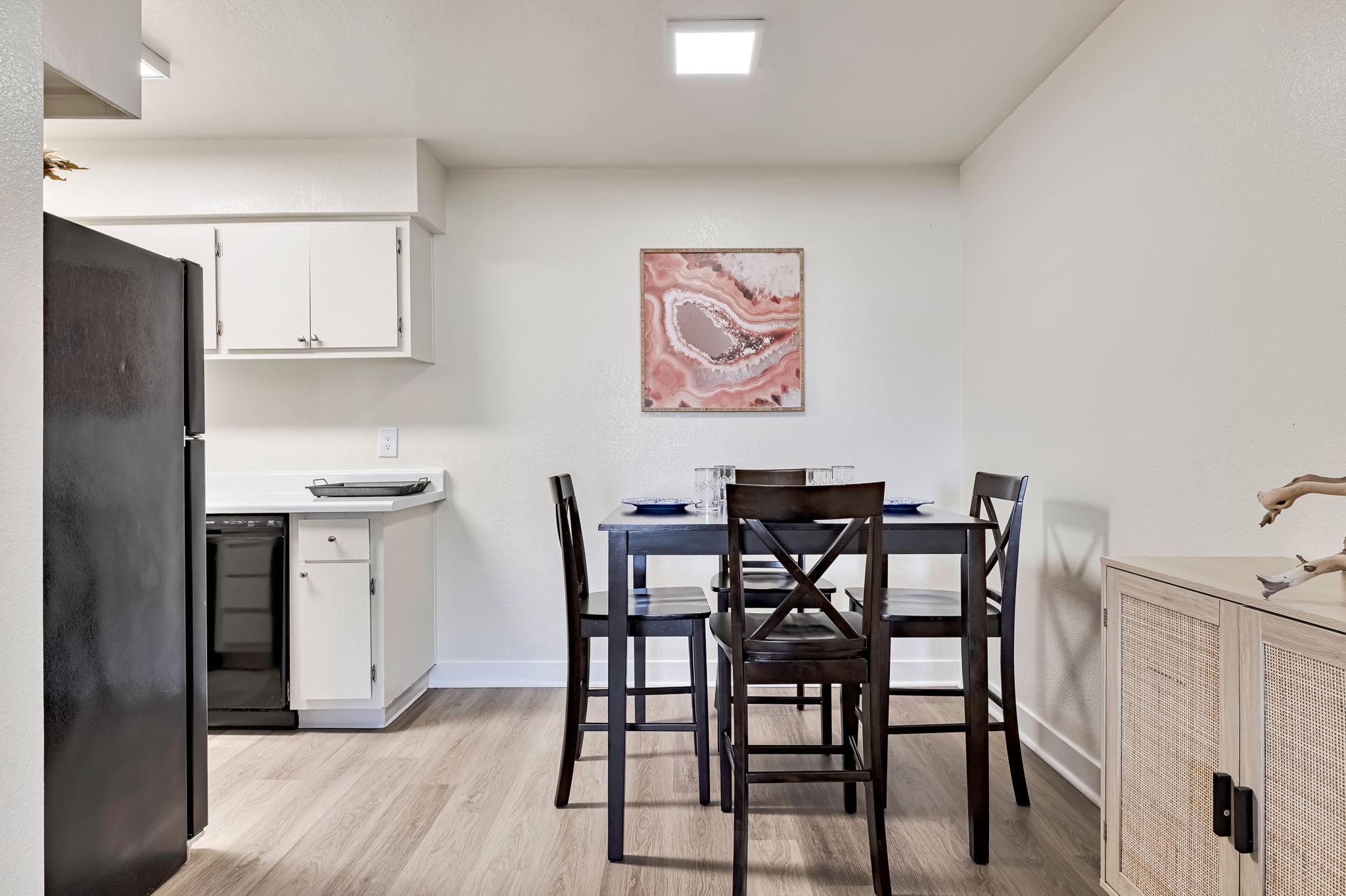 A modern kitchen and dining area featuring a black refrigerator, white cabinets, and a small dining table with four chairs. A piece of abstract art in soft pinks and whites hangs on the wall. The space has light-colored flooring and minimalist decor, creating a bright and inviting atmosphere.