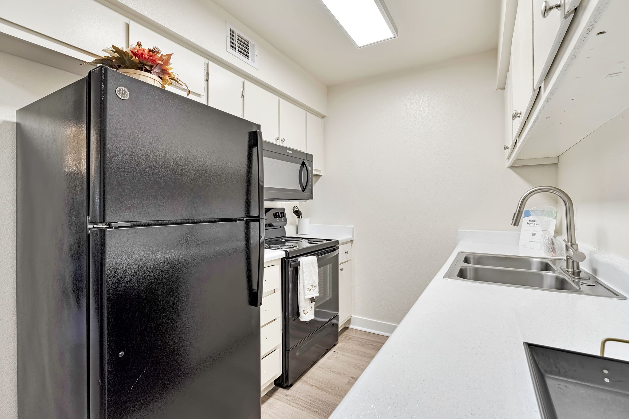 Modern kitchen featuring a black refrigerator, stainless steel microwave, and an electric stove. Light-colored cabinets and countertops create a clean look, with a double sink and neutral walls. A decorative arrangement adds a touch of color on top of the refrigerator.