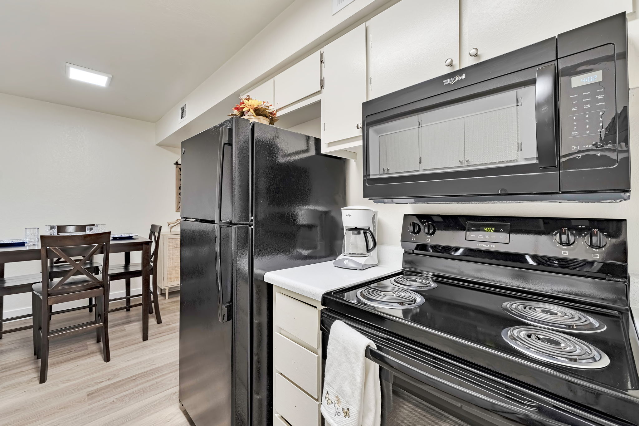 A modern kitchen featuring a black fridge and microwave, an electric stove with an oven, and a coffee maker. There's a small dining table with high chairs in the background, and the floor is covered with light-colored wood laminate. The cabinets are white with simple hardware.