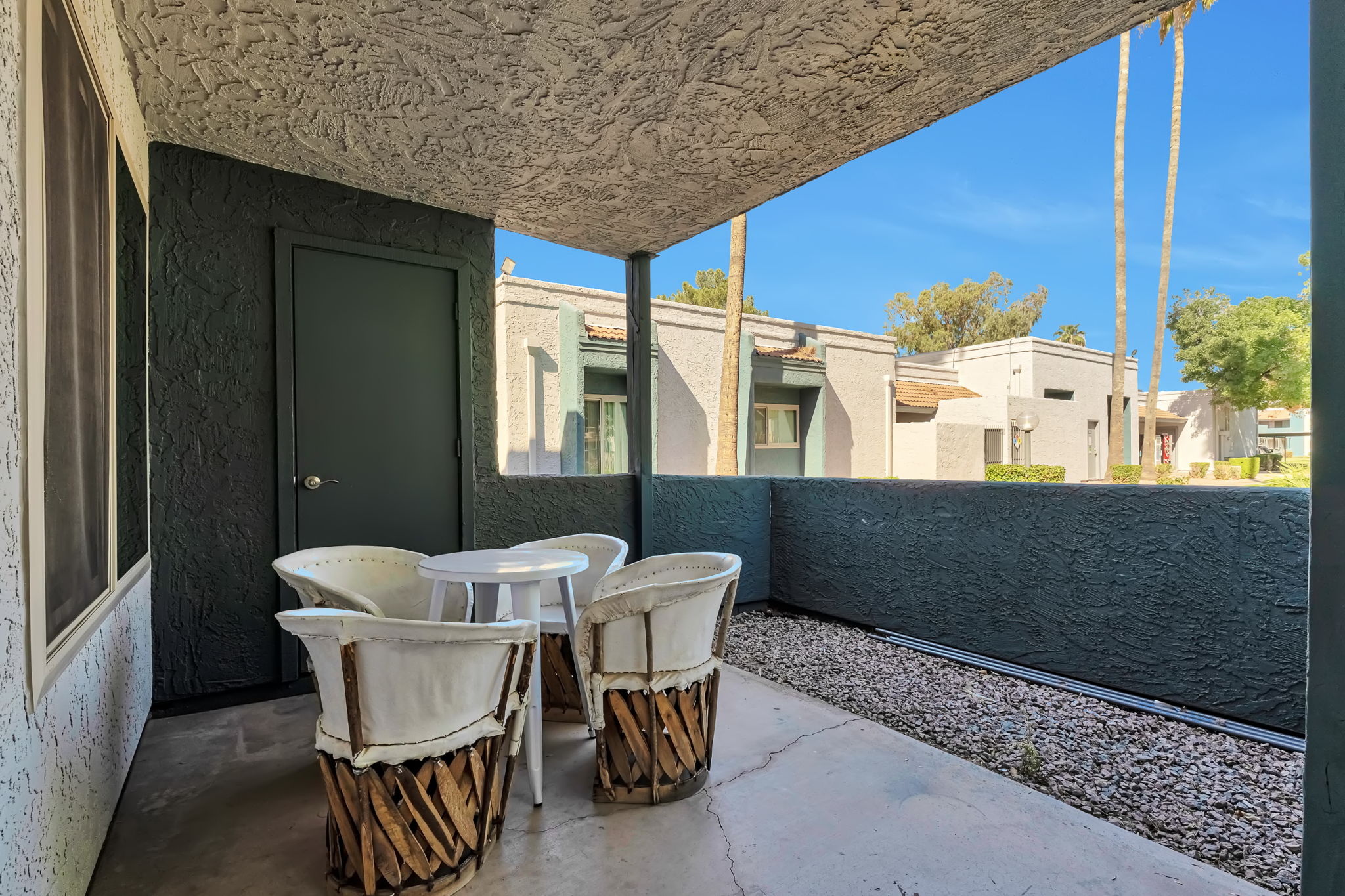 A small outdoor patio with a round white table and four white chairs featuring wooden bases. The setting is surrounded by gray wall textures, and in the background, there are palm trees and neighboring buildings under a clear blue sky.