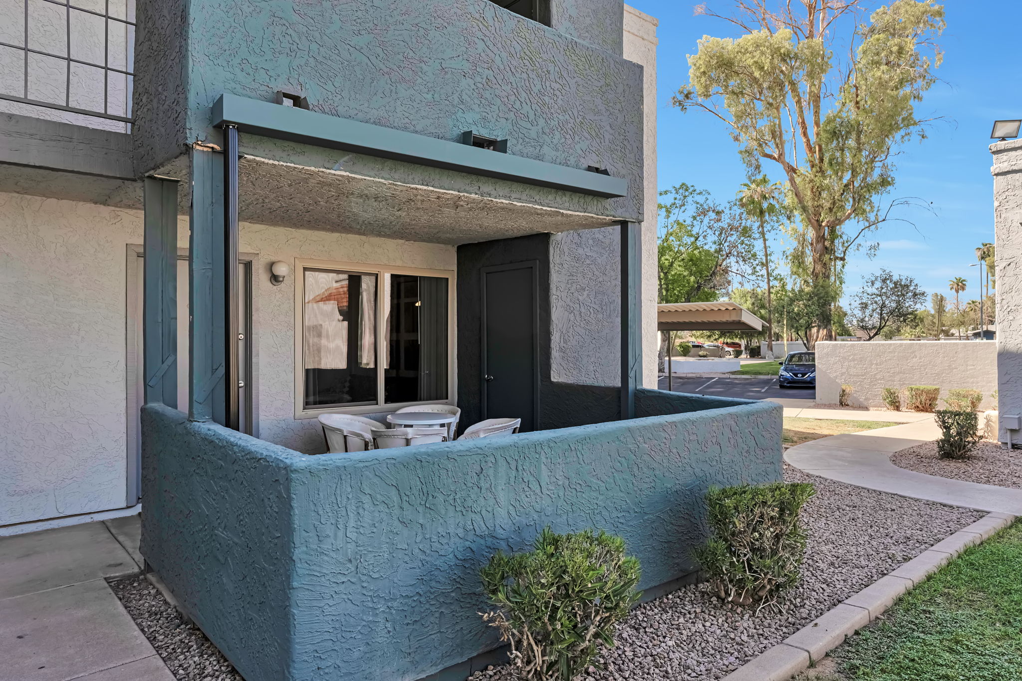 A modern apartment balcony with a covered patio area, featuring a table and chairs. Surrounding landscaping includes small shrubs and gravel. The building has a textured exterior and is in a residential neighborhood with clear skies and nearby parked cars.