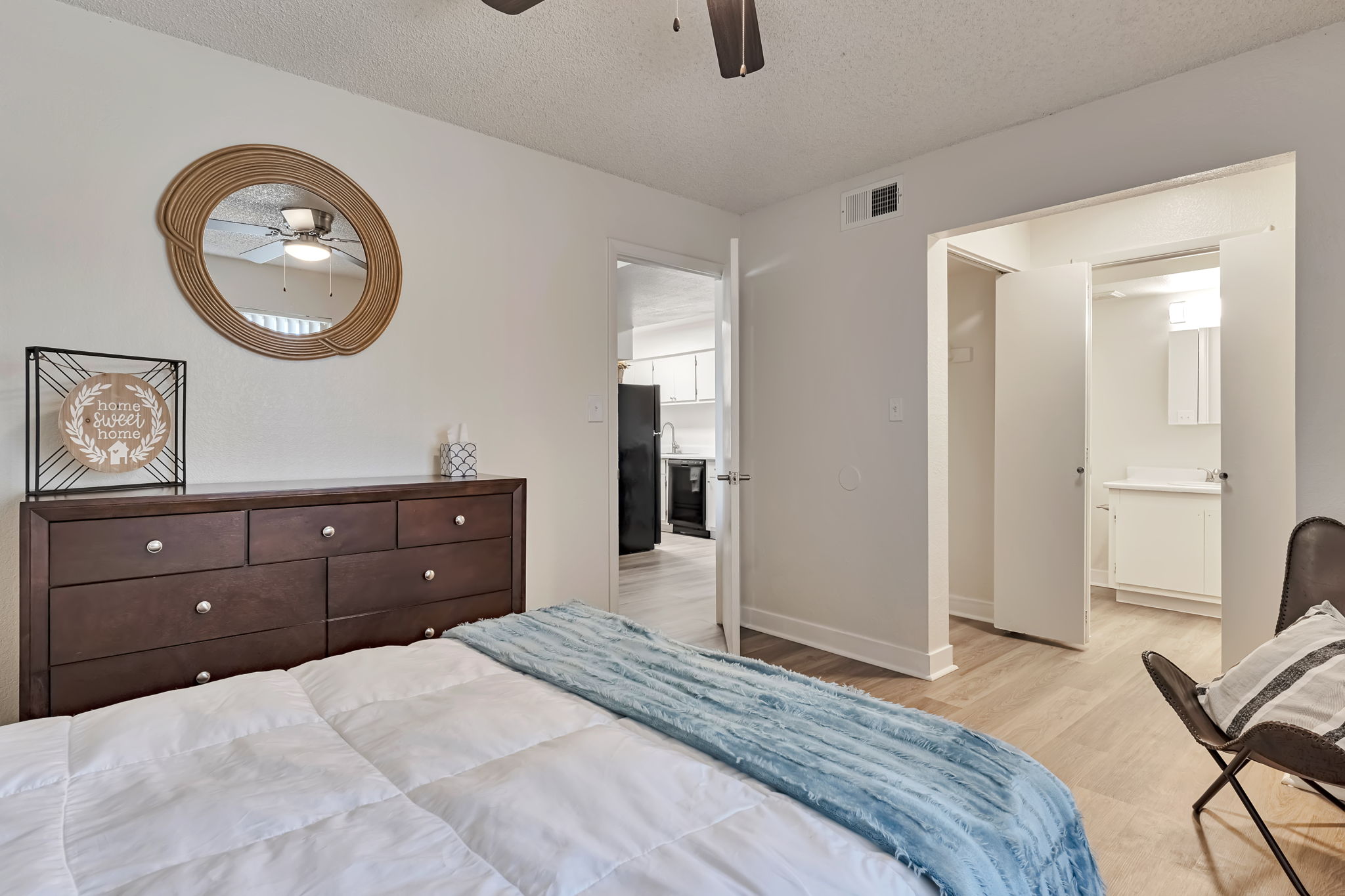 A cozy bedroom featuring a brown dresser with decorative items, a round mirror on the wall, and a bed with a light blue blanket. A doorway leads to a bathroom, and the decor is modern and inviting, with light wood flooring and neutral wall colors.