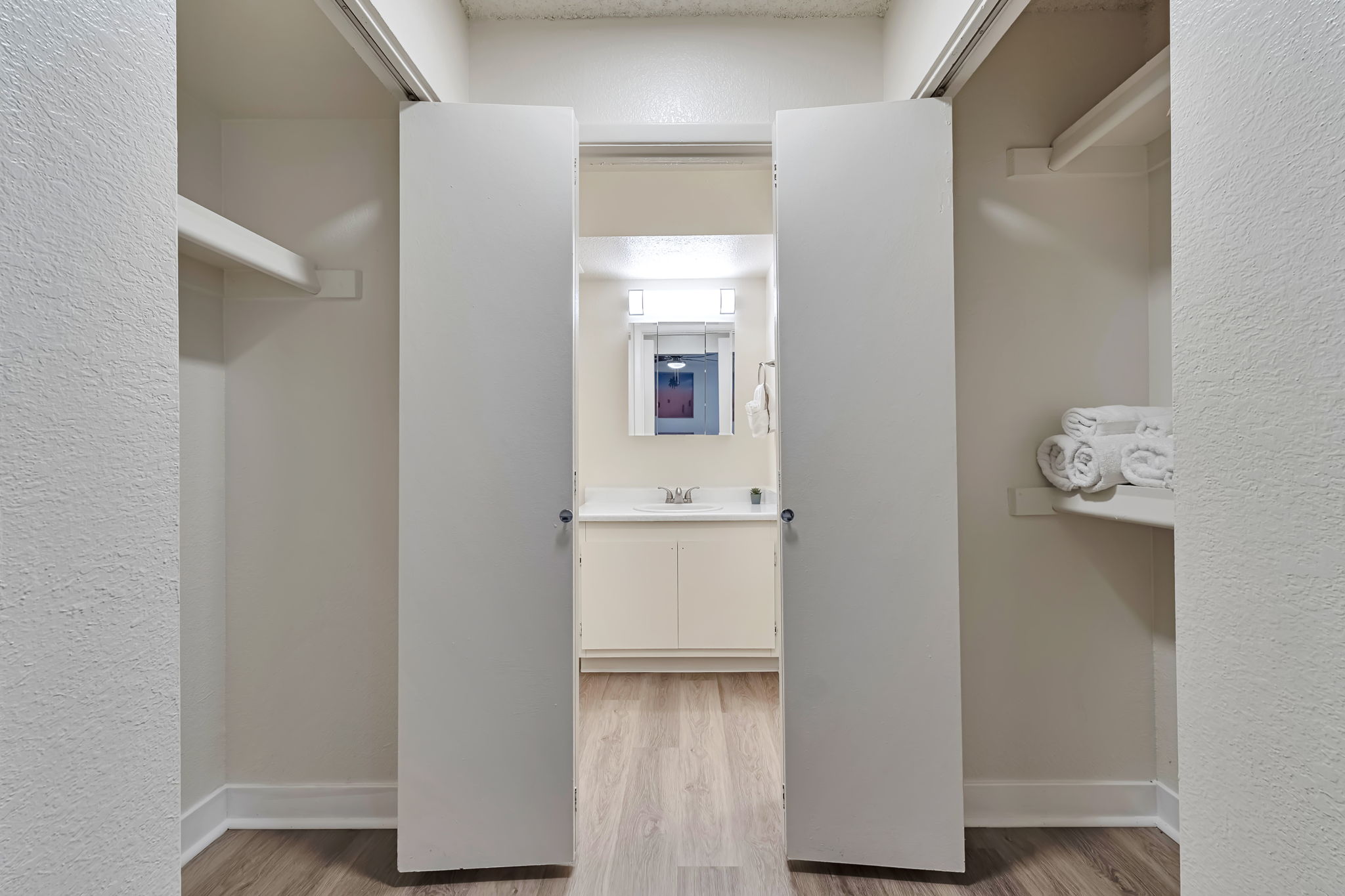 Interior view of a closet with open double doors leading to a bright bathroom. The bathroom features a mirror above a sink and neatly stacked white towels on a shelf. The walls and flooring are light-colored, creating an airy and clean atmosphere.