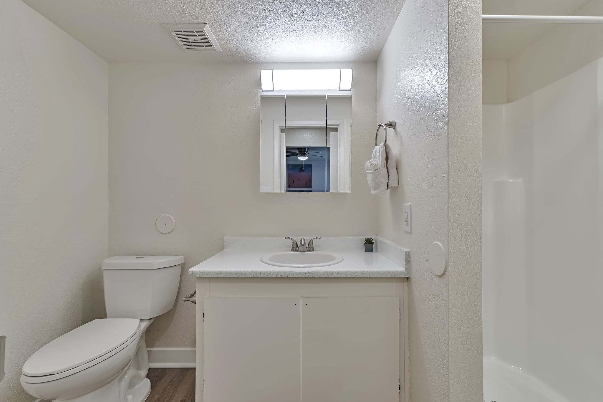 A clean, modern bathroom featuring a white vanity with a round sink, a toilet, and a standing shower stall. The walls are painted a neutral color, and there is a small window above the sink with natural light. A towel hangs on a rack, and a small plant is on the countertop.