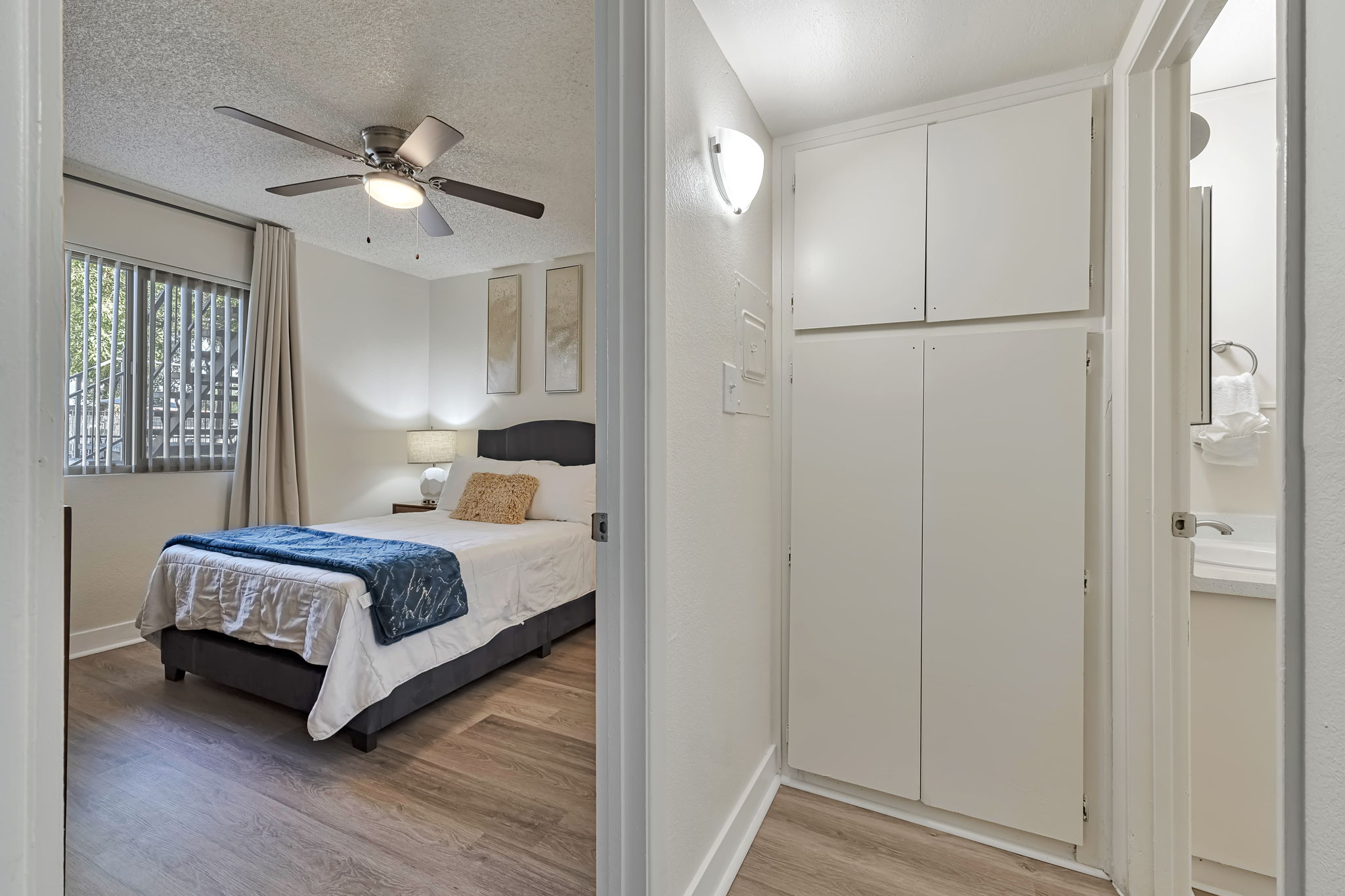 A bright and airy hotel room featuring a bed with white bedding and a decorative blue throw. A ceiling fan is above, and the room is decorated with wall art. A small hallway leads to a bathroom area with a mirrored sink and storage cabinets, with natural light coming from a window in the background.