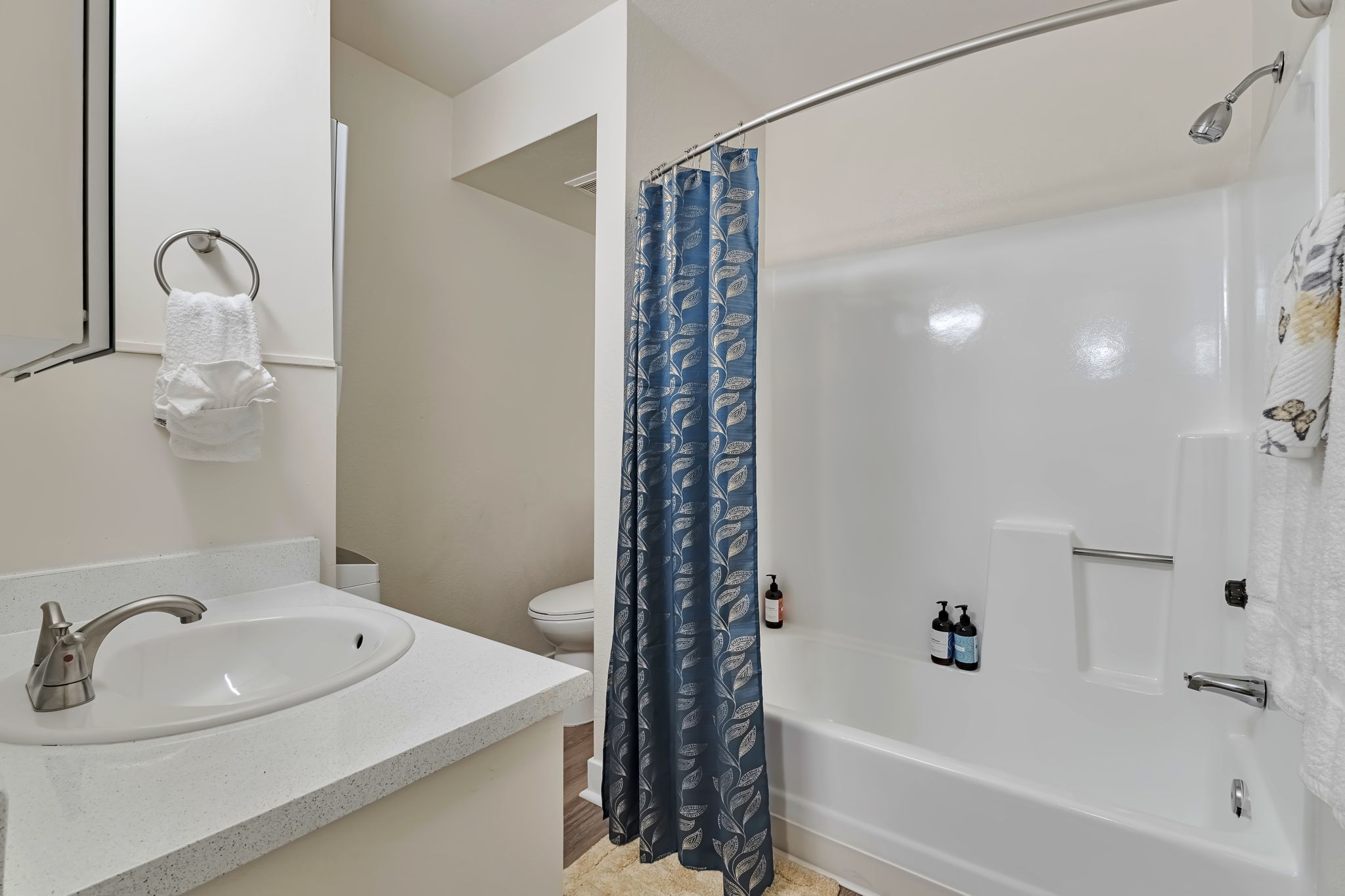 A clean and modern bathroom featuring a white bathtub with a shower curtain, a minimalist sink with a faucet, and toiletries on the edge of the tub. Light-colored walls and a towel rack with neatly hung towels contribute to a tidy appearance. A toilet is visible in the background.