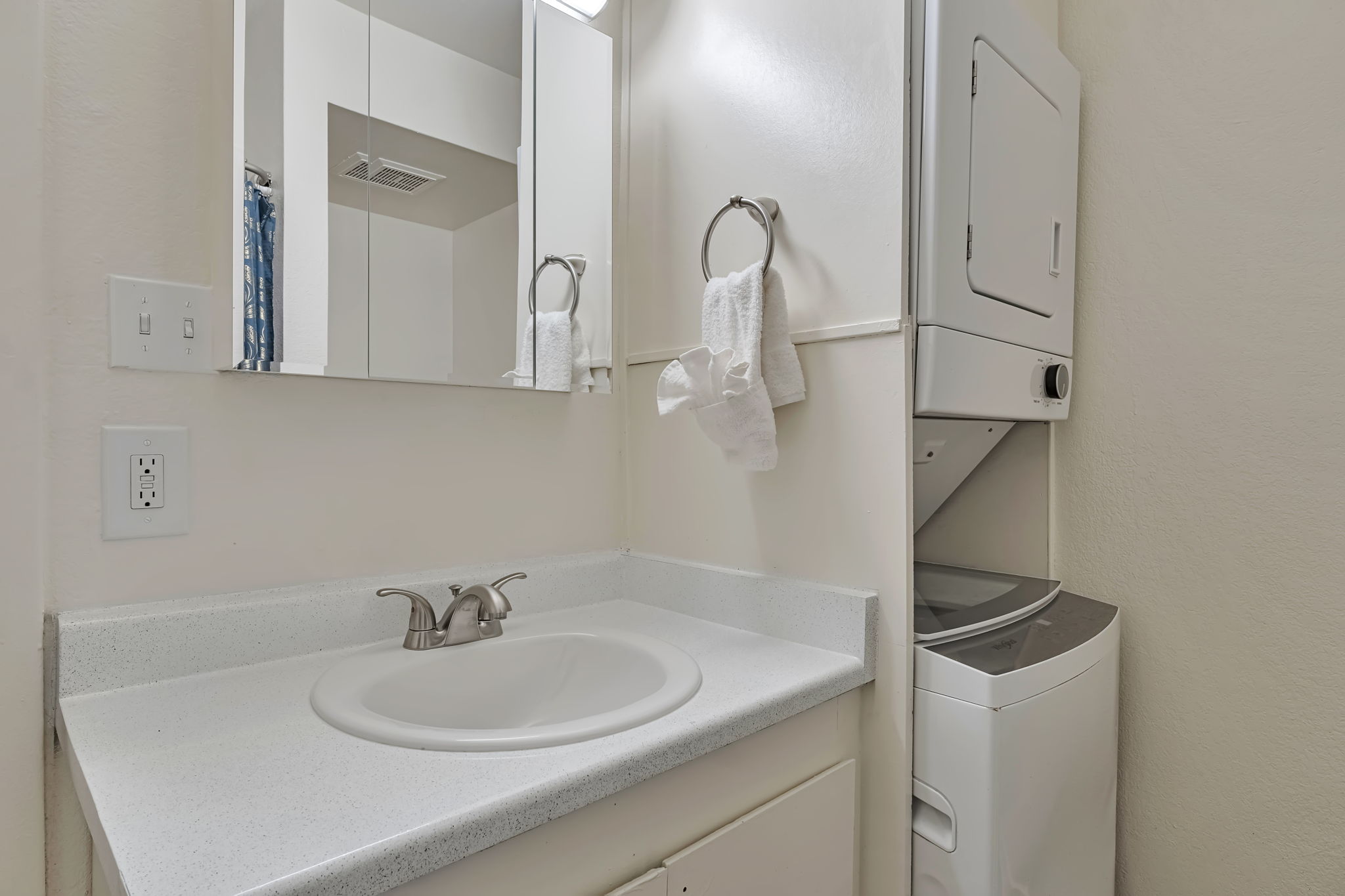 Small bathroom featuring a white sink with a faucet, a wall-mounted mirror, and two white towels hanging on a towel bar. To the right, there is a stacked washer and dryer unit, and the walls are painted in a light color. The space is bright and clean.
