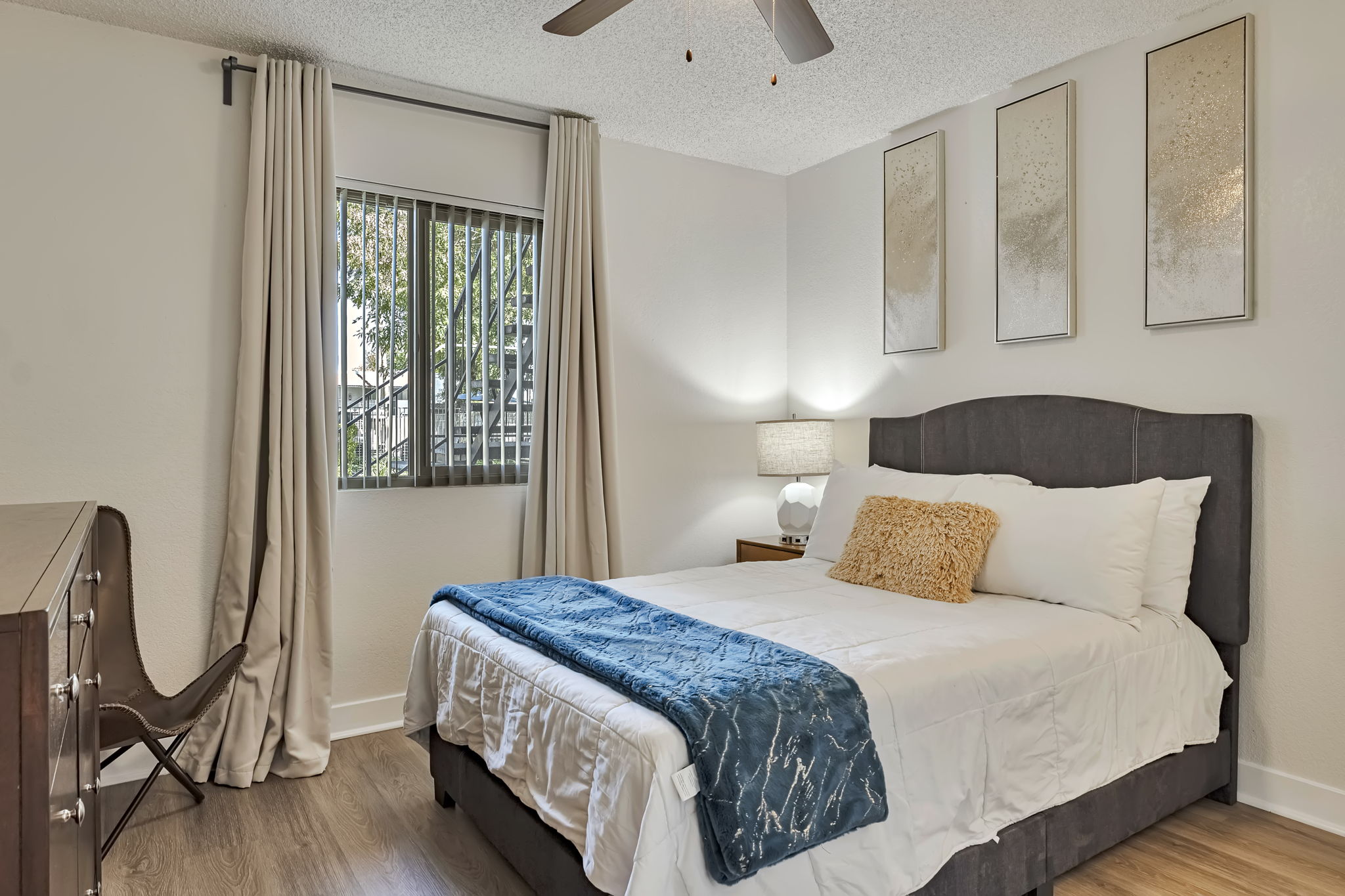 A modern bedroom featuring a queen-sized bed with white bedding and a decorative blue throw. Natural light filters through a window with vertical blinds. A nightstand with a lamp sits beside the bed, and three framed artworks adorn the wall. The room has wood flooring and a stylish chair in the corner.