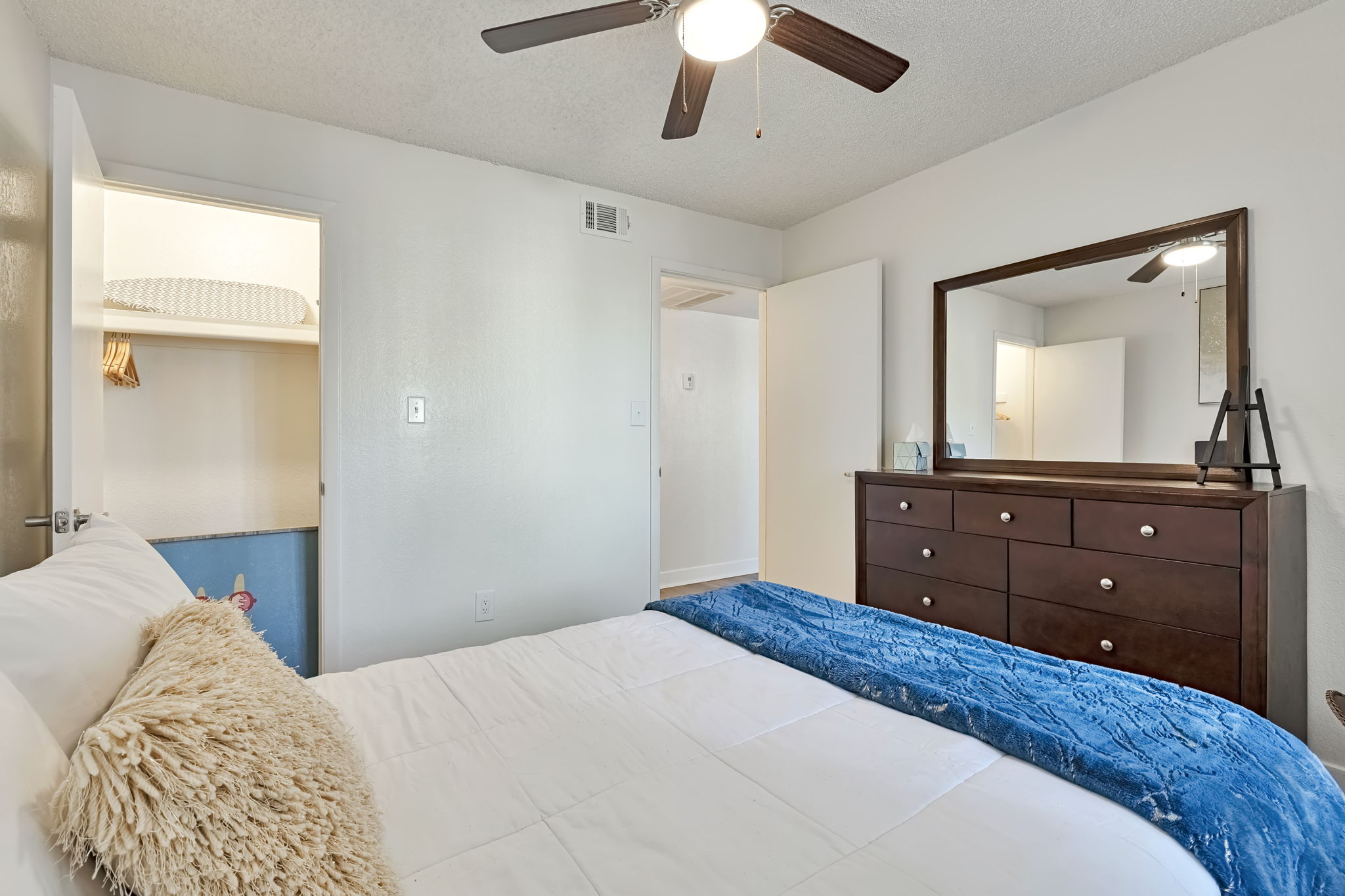 A cozy bedroom featuring a plush bed with a decorative pillow, a wooden dresser, and a mirror. A ceiling fan is above, and there's a closet visible through an open door in the background. The walls are painted light colors, contributing to a bright and inviting atmosphere.