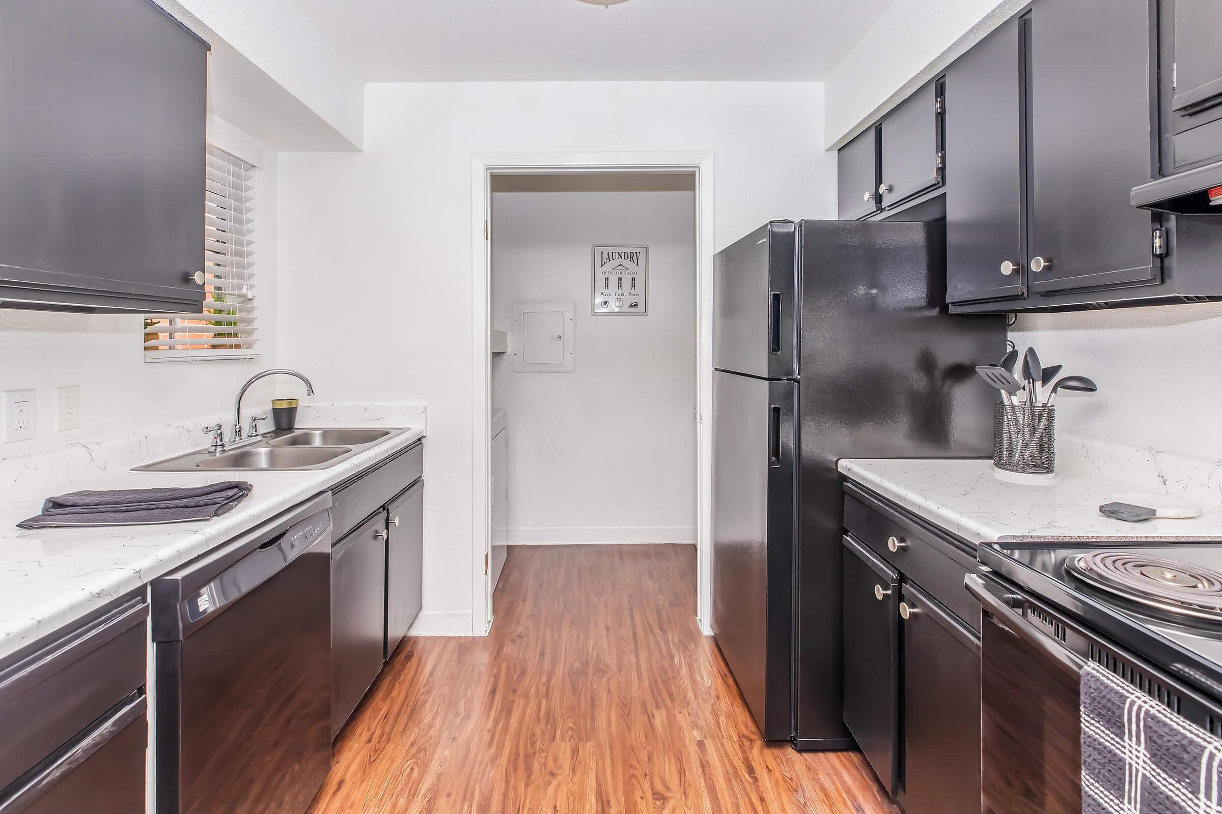 A modern kitchen featuring dark cabinets, a double sink, and a black refrigerator. The countertops are white with a marbled design. Appliances include a stove, microwave, and utensil holder. The floor is wood laminate, and there is a doorway leading to an adjoining room. Natural light comes through a window.