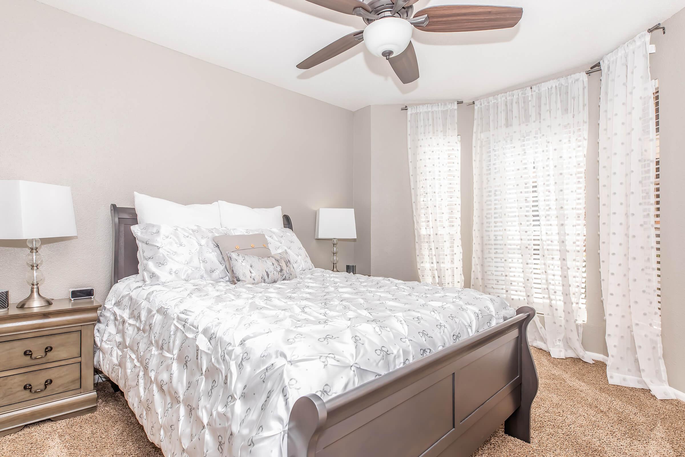 A cozy bedroom featuring a large bed with white bedding and decorative pillows. The room includes two bedside lamps on wooden nightstands and a ceiling fan. Bright curtains frame the windows, allowing natural light to fill the space, which has plush carpeting and neutral-colored walls.