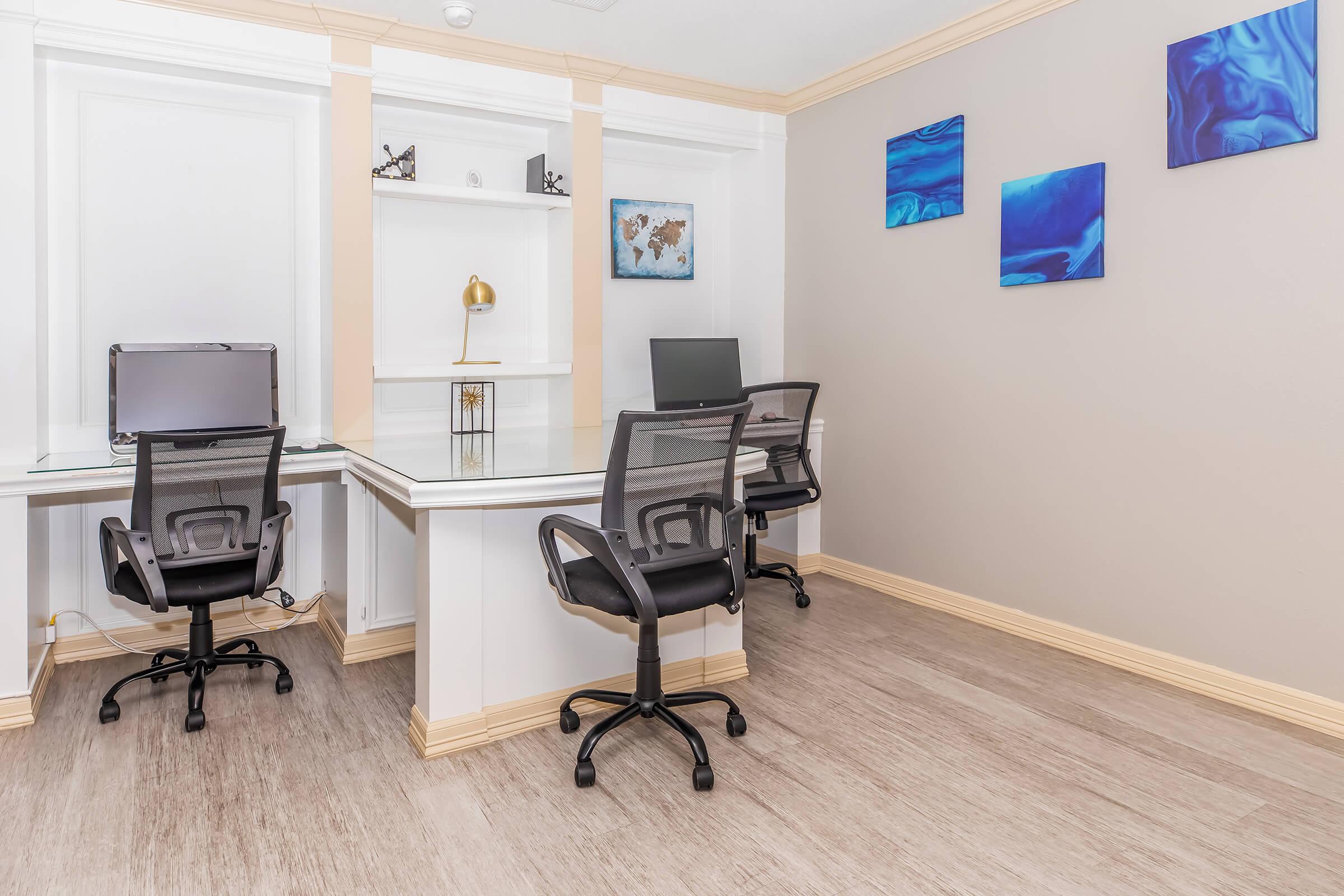 A modern office space featuring two sleek black office chairs at glass desks. One desk has a computer monitor, while the other is empty. The room is decorated with blue abstract artwork on the walls, and it has light-colored wooden flooring and beige walls for a clean, professional look.
