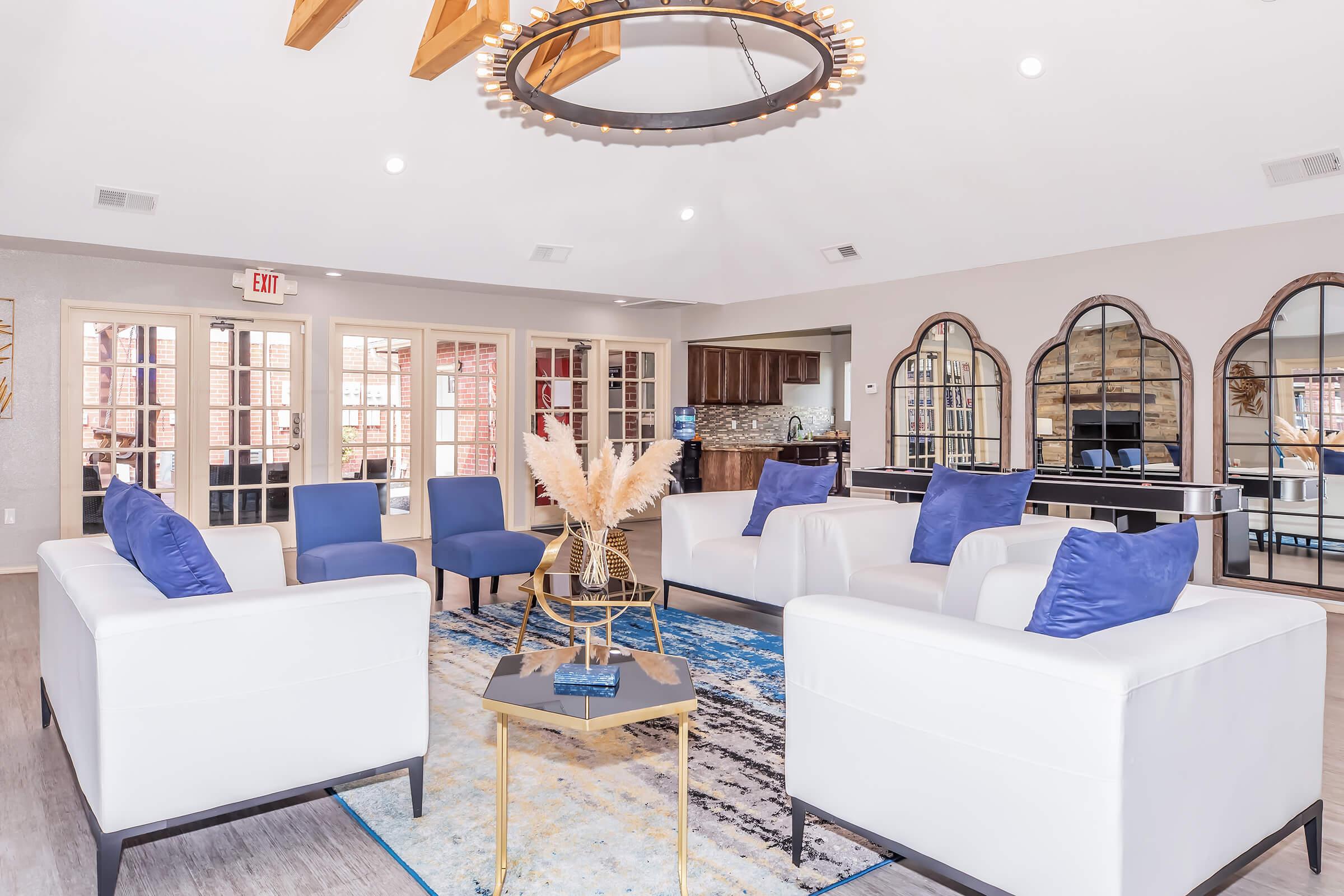 A modern lounge area featuring white sofas with blue cushions, blue chairs, a round coffee table, and decorative plants. Large windows let in natural light, and the space includes stylish mirrors and a shiny chandelier. The walls are painted in neutral tones, complementing the contemporary décor.