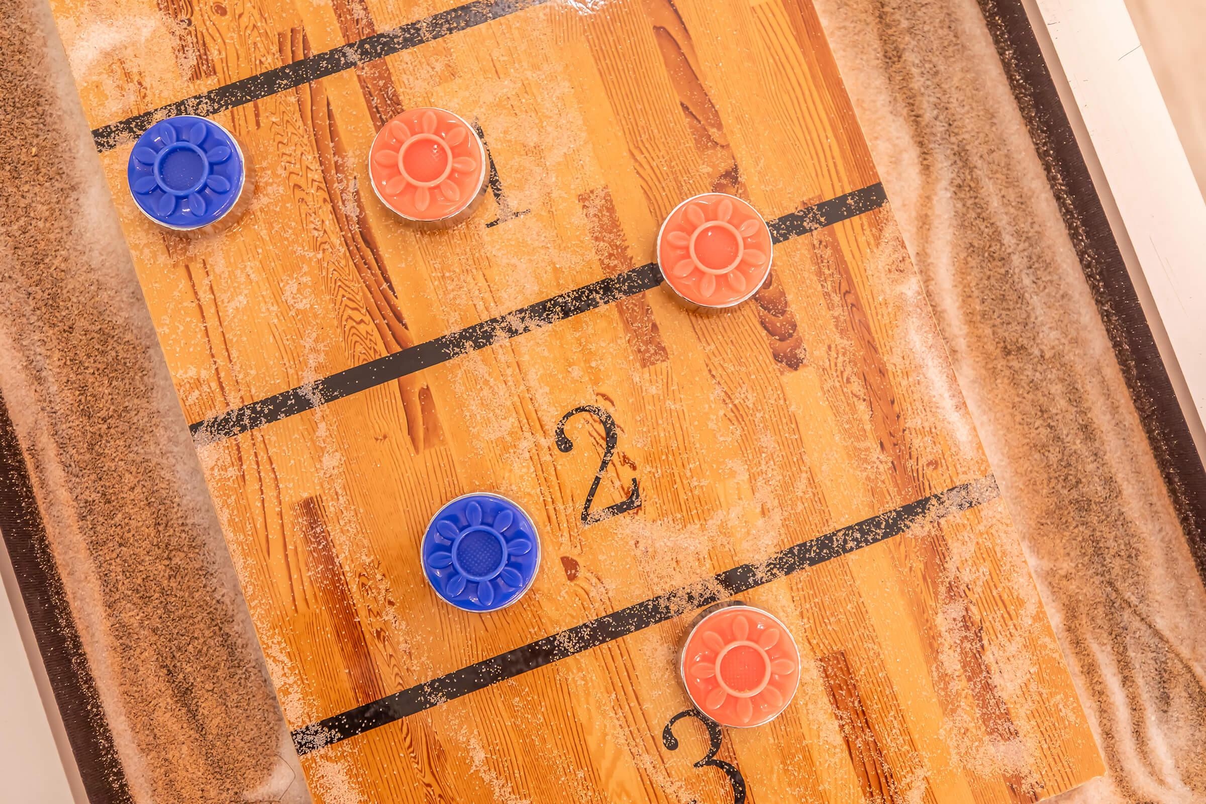 A close-up view of a wooden shuffleboard table with a sandy playing surface. Two orange and two blue pucks are positioned on the table, with numbers painted on the surface indicating scoring zones. The background shows a wooden border around the shuffleboard area.
