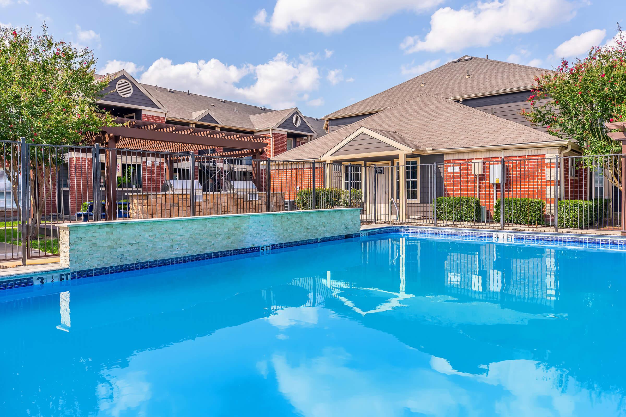 A clear blue swimming pool with a stone wall feature is in the foreground, surrounded by a fence. In the background, there are two residential buildings with a pergola and seating area, under a bright blue sky with scattered clouds. Lush green trees frame the scene.