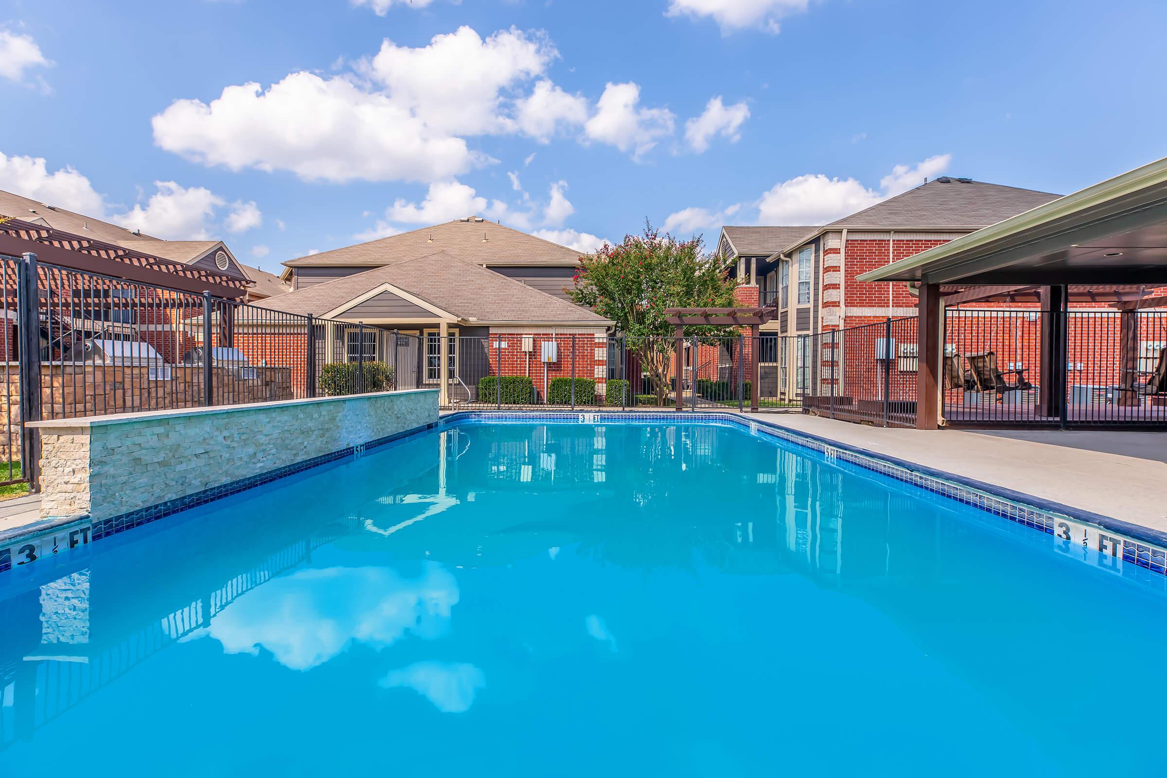 A clear blue swimming pool surrounded by a wrought iron fence, with several apartment buildings in the background. The sky is bright with fluffy clouds, and there are shaded areas with pergolas and greenery nearby. The scene suggests a relaxing outdoor space for residents.