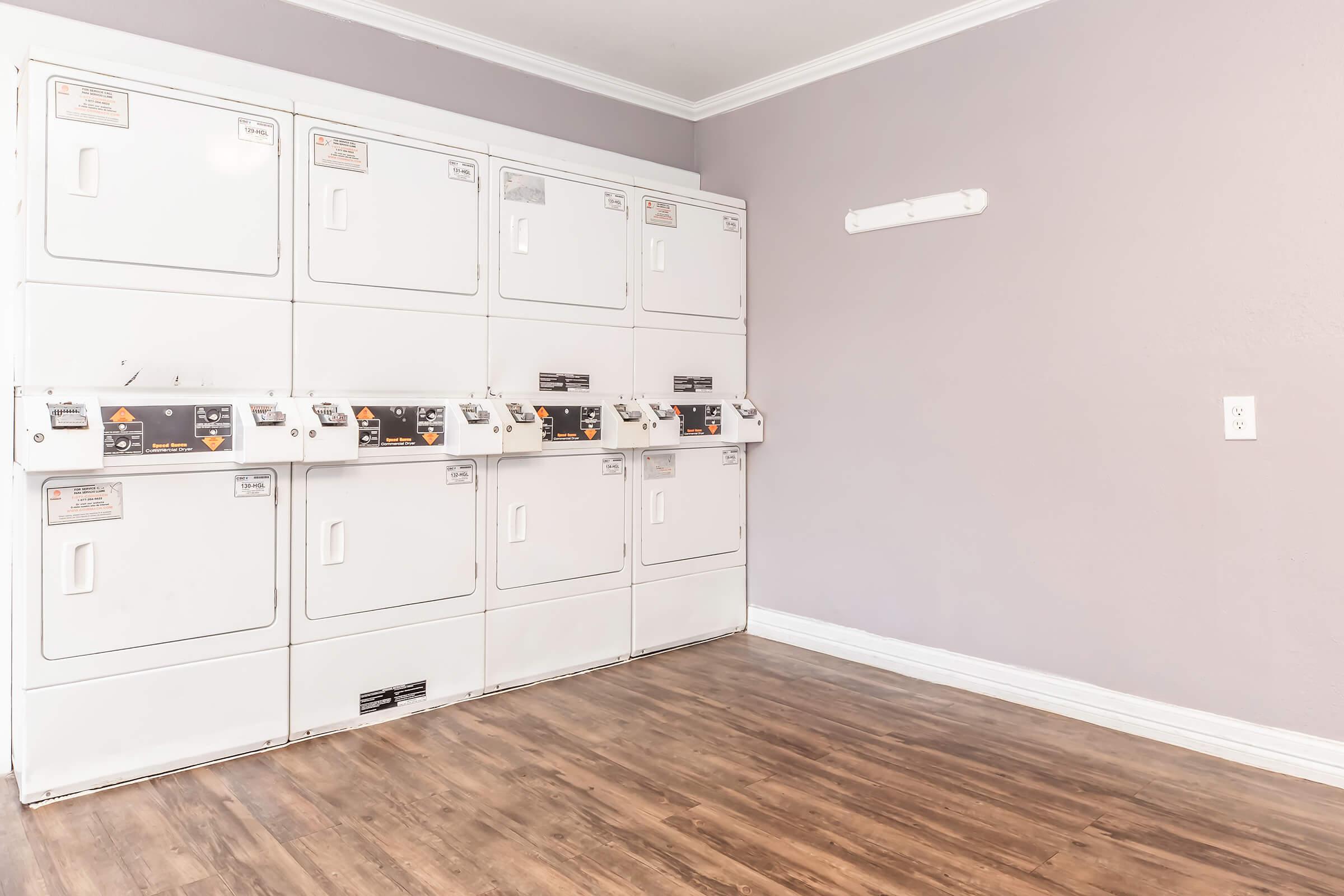 A modern laundry room featuring several stacked white washing machines and dryers against a light purple wall. The flooring is a warm wood finish, and there is a light fixture on the wall. The space is clean and uncluttered, ideal for laundry activities.