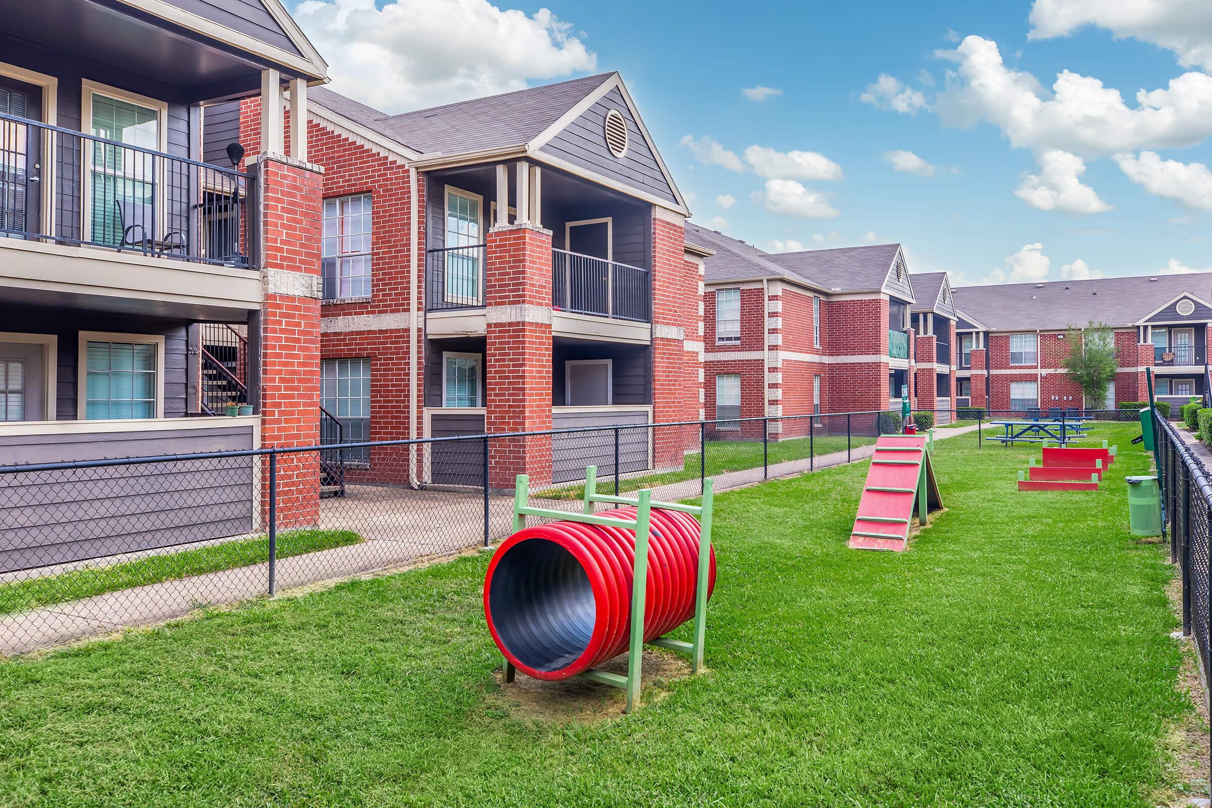 A well-maintained outdoor playground area featuring a red tunnel slide and a small climbing structure on lush green grass. Surrounding the playground are brick apartment buildings with balconies, enclosed by a chain-link fence under a bright blue sky with fluffy clouds.