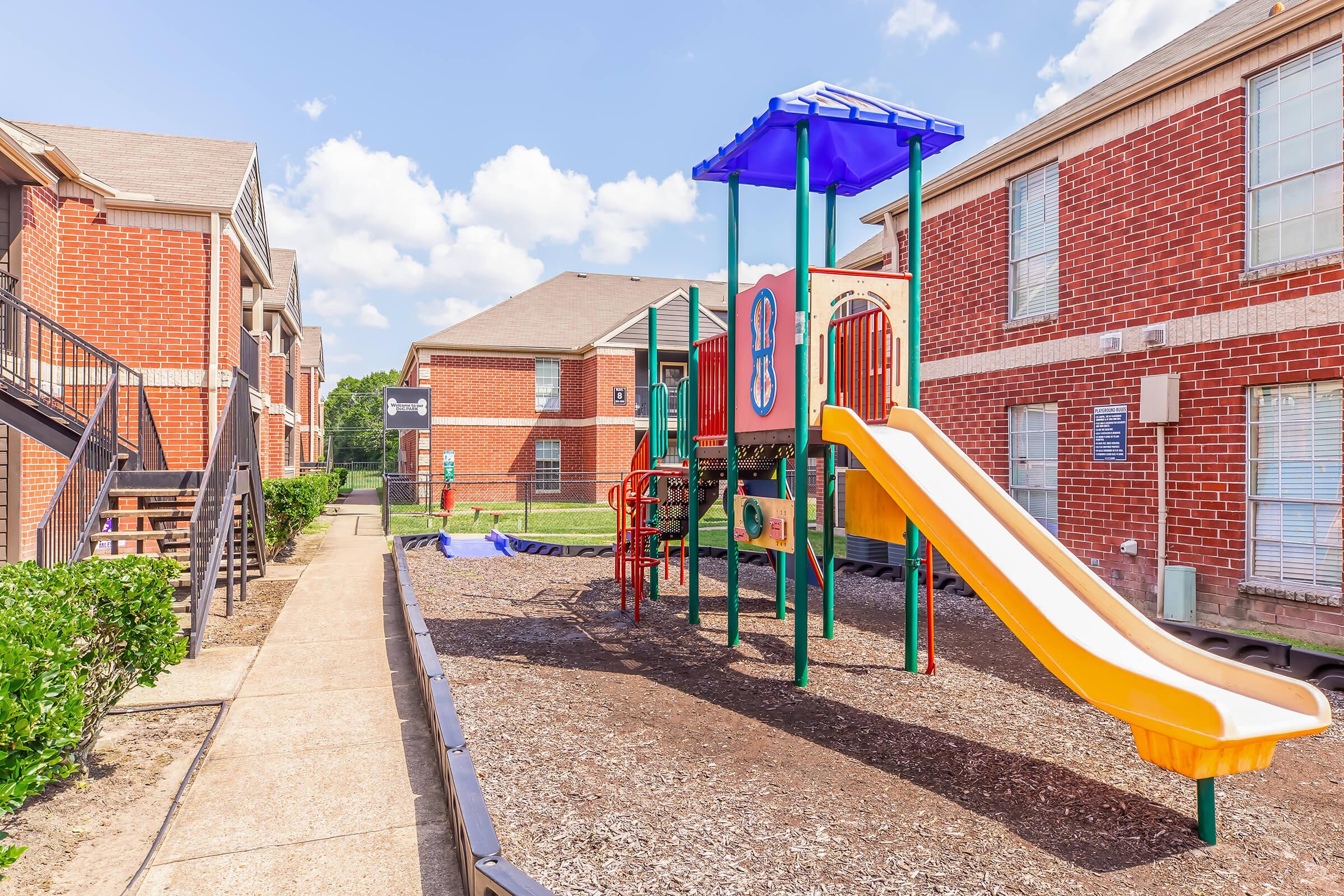 Playground area with a colorful play structure featuring a slide and climbing elements, surrounded by grass and pathways. Red brick apartment buildings in the background under a blue sky with fluffy clouds.
