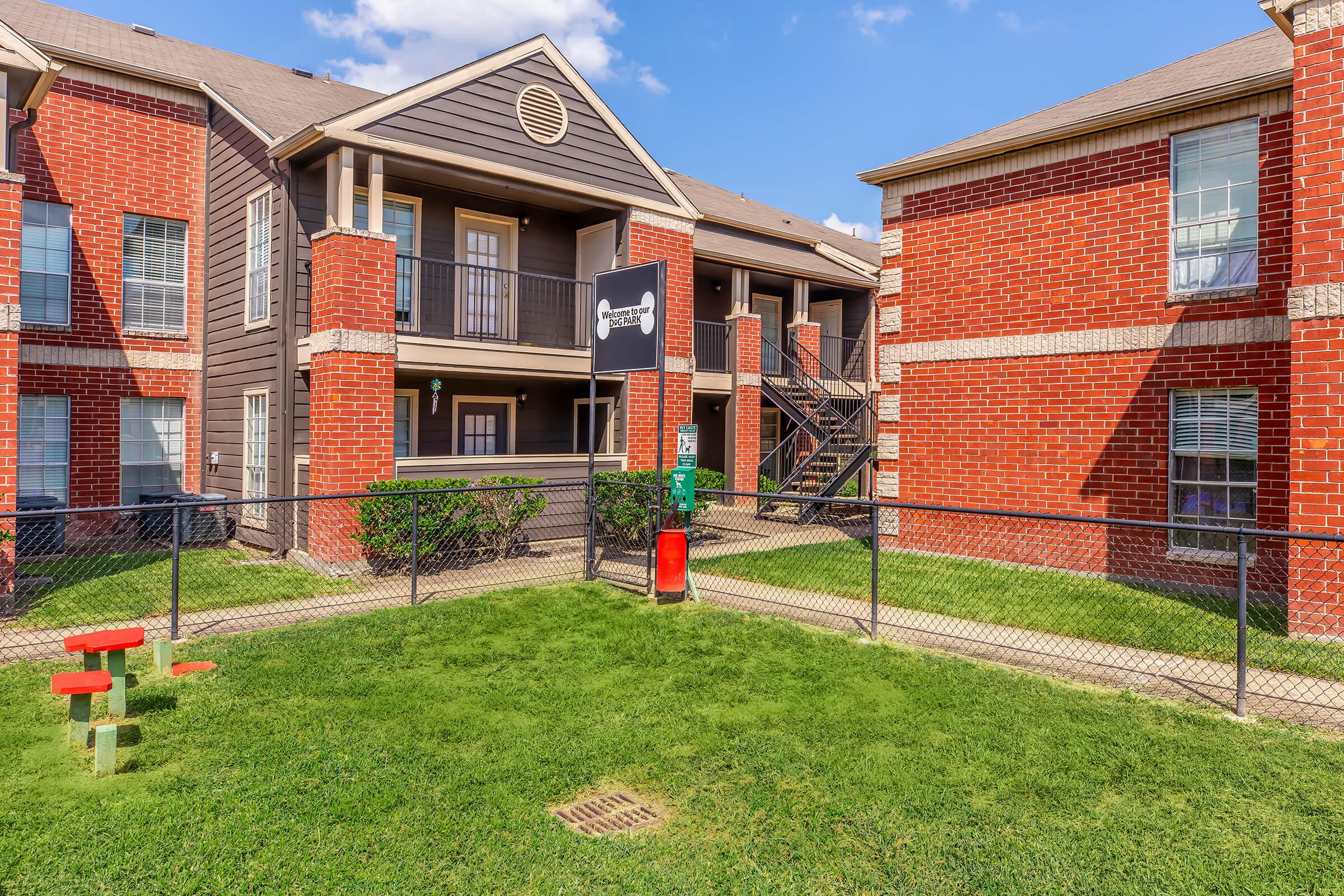 A two-story apartment building with a mix of dark wood and brick exteriors. The ground level features a sign and a green lawn enclosed by a chain-link fence. There are outdoor stairs leading to the upper floor, and colorful playground equipment is visible in the foreground. Bright blue sky adds to the cheerful atmosphere.