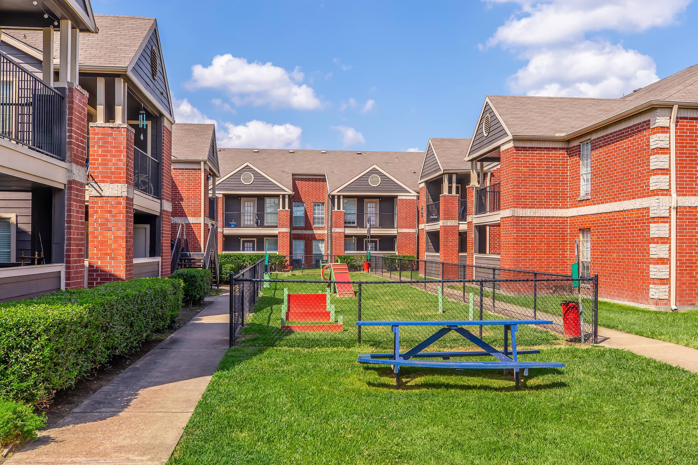 A peaceful courtyard surrounded by brick apartment buildings. The area features a well-maintained green lawn, a picnic table, and red steps leading to a central play area, all under a bright blue sky with fluffy white clouds. The scene conveys a welcoming and community-friendly atmosphere.