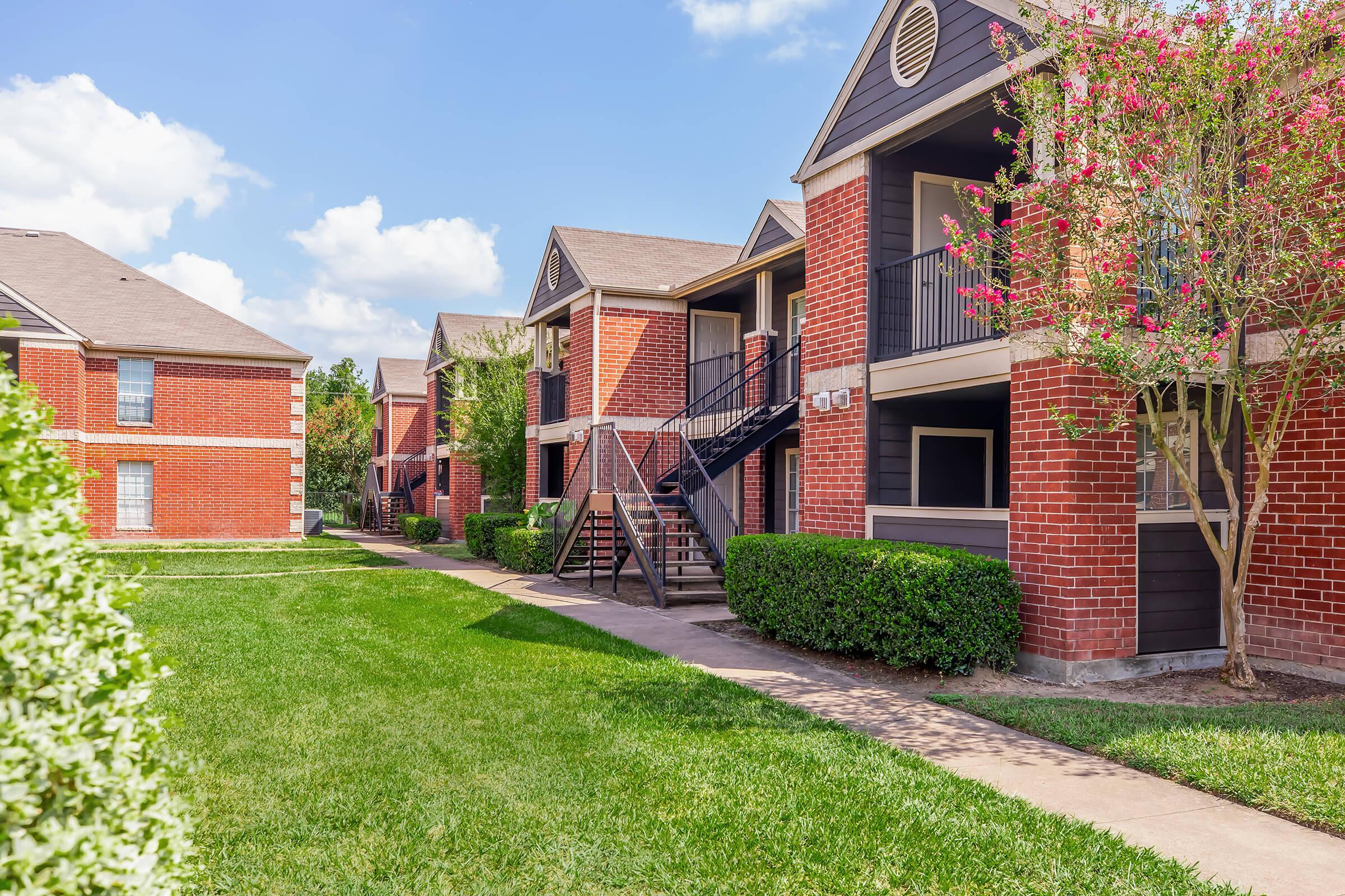 Two-story brick apartment buildings line a manicured pathway with green lawns and shrubs. The buildings feature balconies and black iron railings. The scene is set under a bright blue sky with fluffy white clouds, creating a sunny and inviting atmosphere.