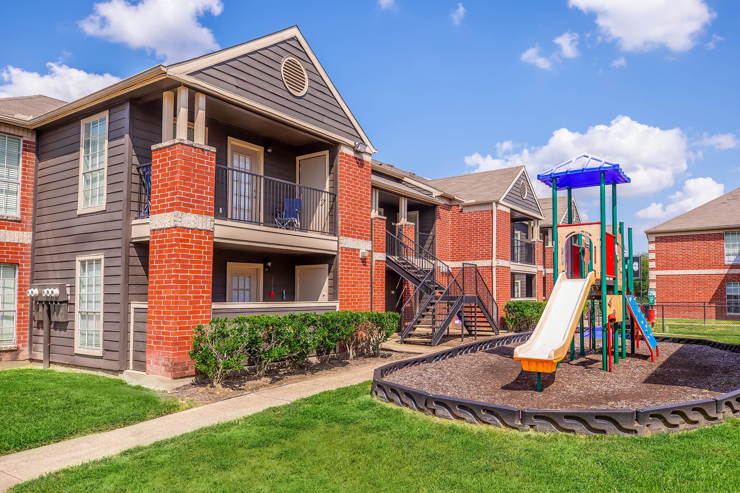 A two-story apartment building featuring a red brick facade and balconies, with a playground including a slide and climbing structure in the foreground. Lush green grass surrounds the area, under a clear blue sky with fluffy white clouds.