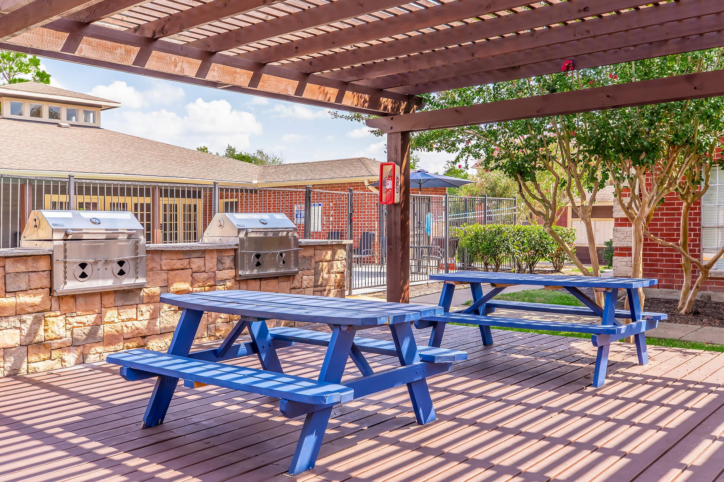 A shaded outdoor area featuring blue picnic tables beneath a wooden pergola. The ground is a wooden deck with shadows cast by the slats above. In the background, there are two metal grills and a well-maintained green space, along with buildings visible beyond a fence.